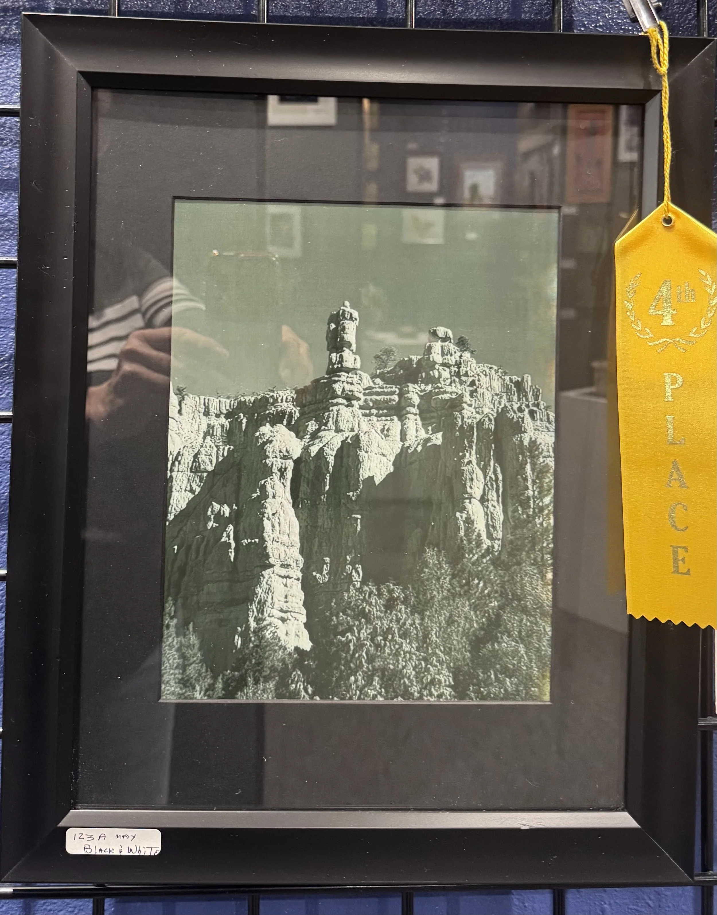 Black picture frame with a yellow ribbon hanging on the top right side, displaying a black and white photograph of a mountain landscape with rocky formations and sparse trees.