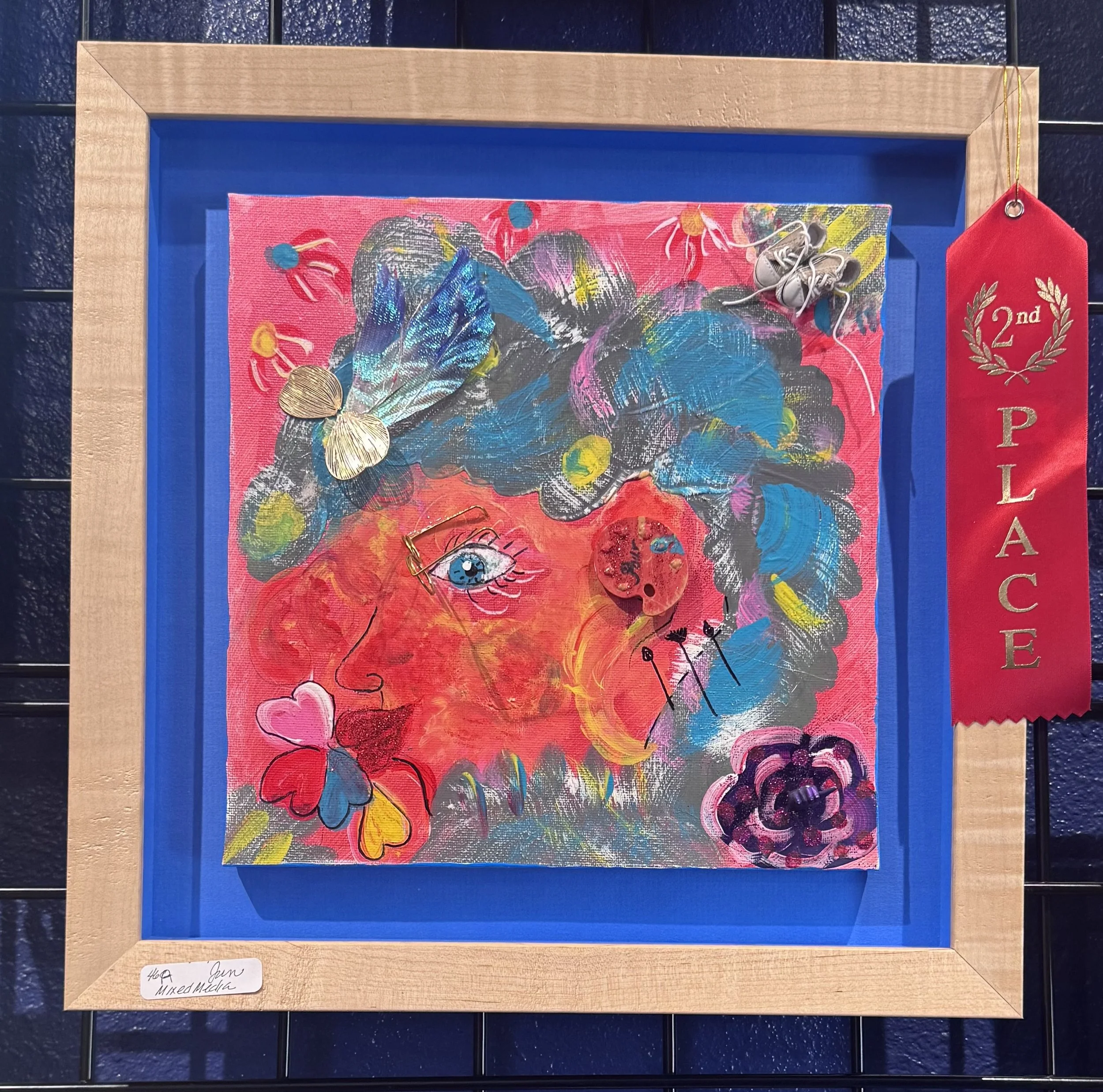 Mixed media artwork featuring colorful abstract shapes, an eye, a butterfly, hearts, and various decorative elements on a pink background, framed in wood with a red ribbon and a pair of miniature shoes attached.
