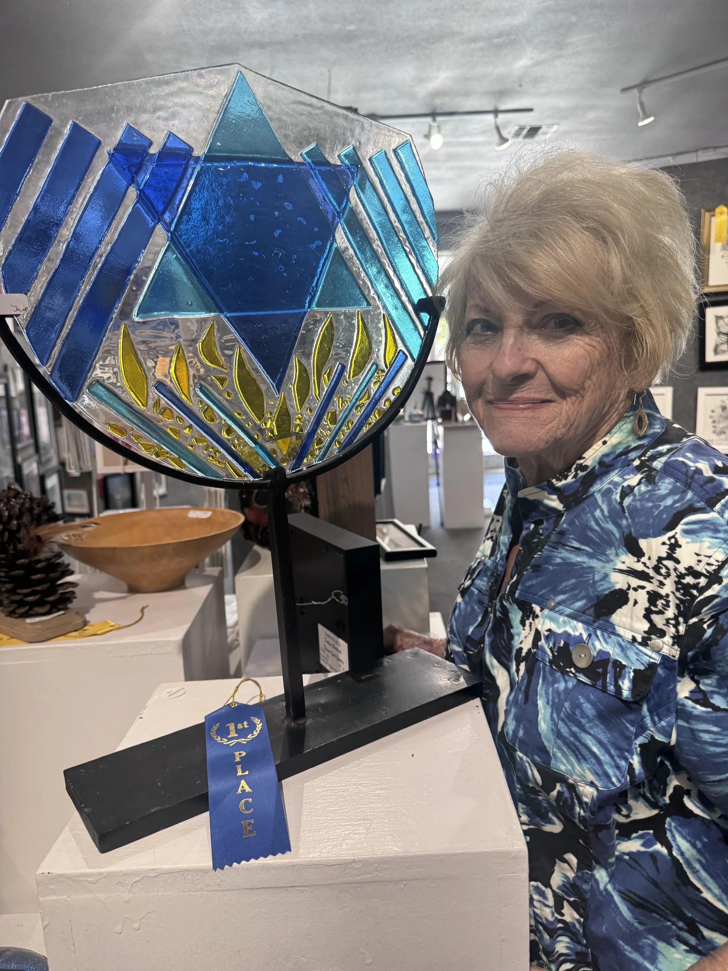 An elderly woman with short, light-colored hair wearing a blue floral shirt stands behind a black stand with a stained glass art piece featuring blue, yellow, and clear glass sections. The art piece has a blue ribbon that says "1st Place." The backgr