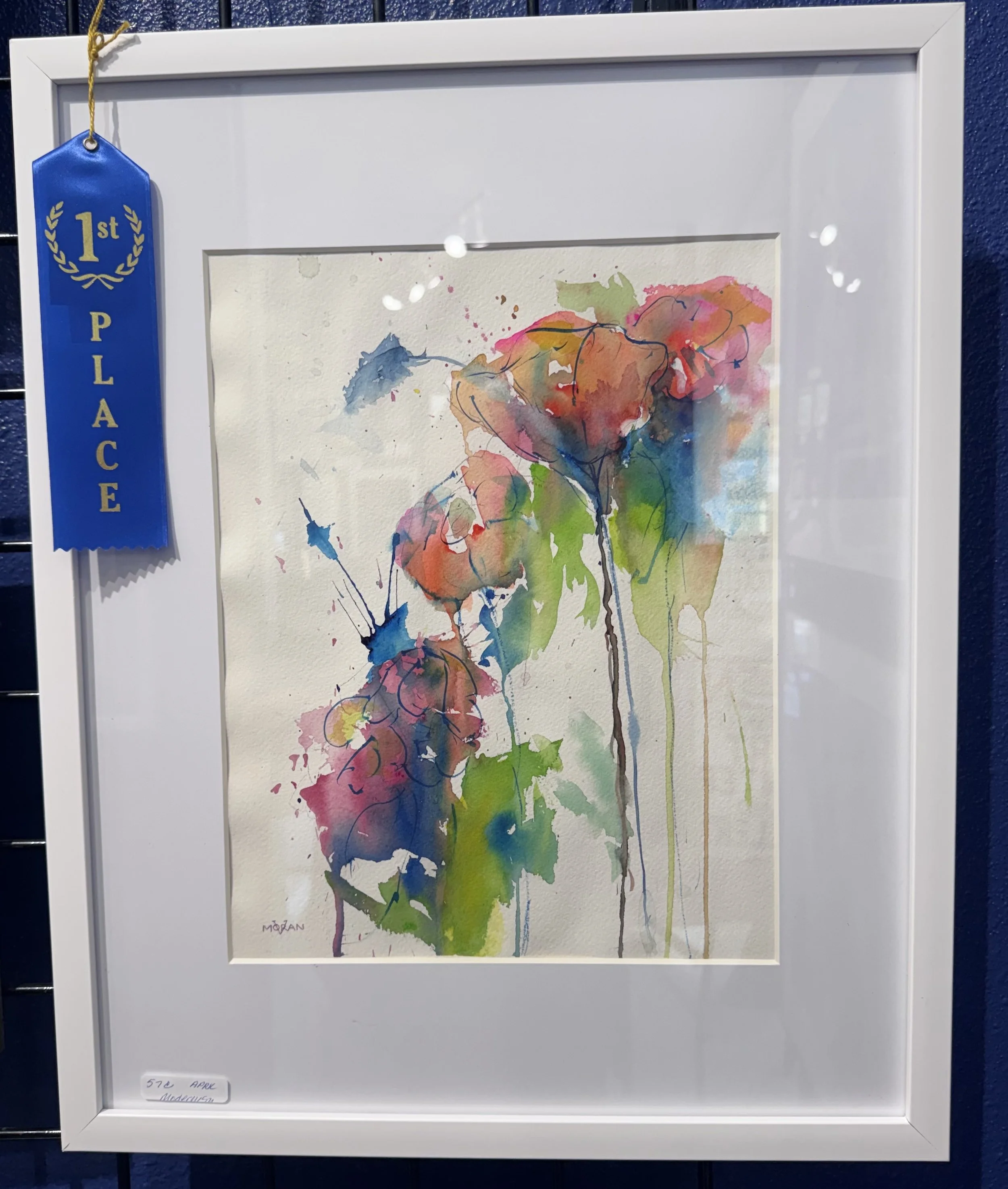 Watercolor painting of trees with leaves in pink, orange, blue, green, and brown on watercolor paper framed with a white mat, hanging on a blue wall. A blue ribbon with gold lettering that says '1st Place' is attached to the top left corner of the fr