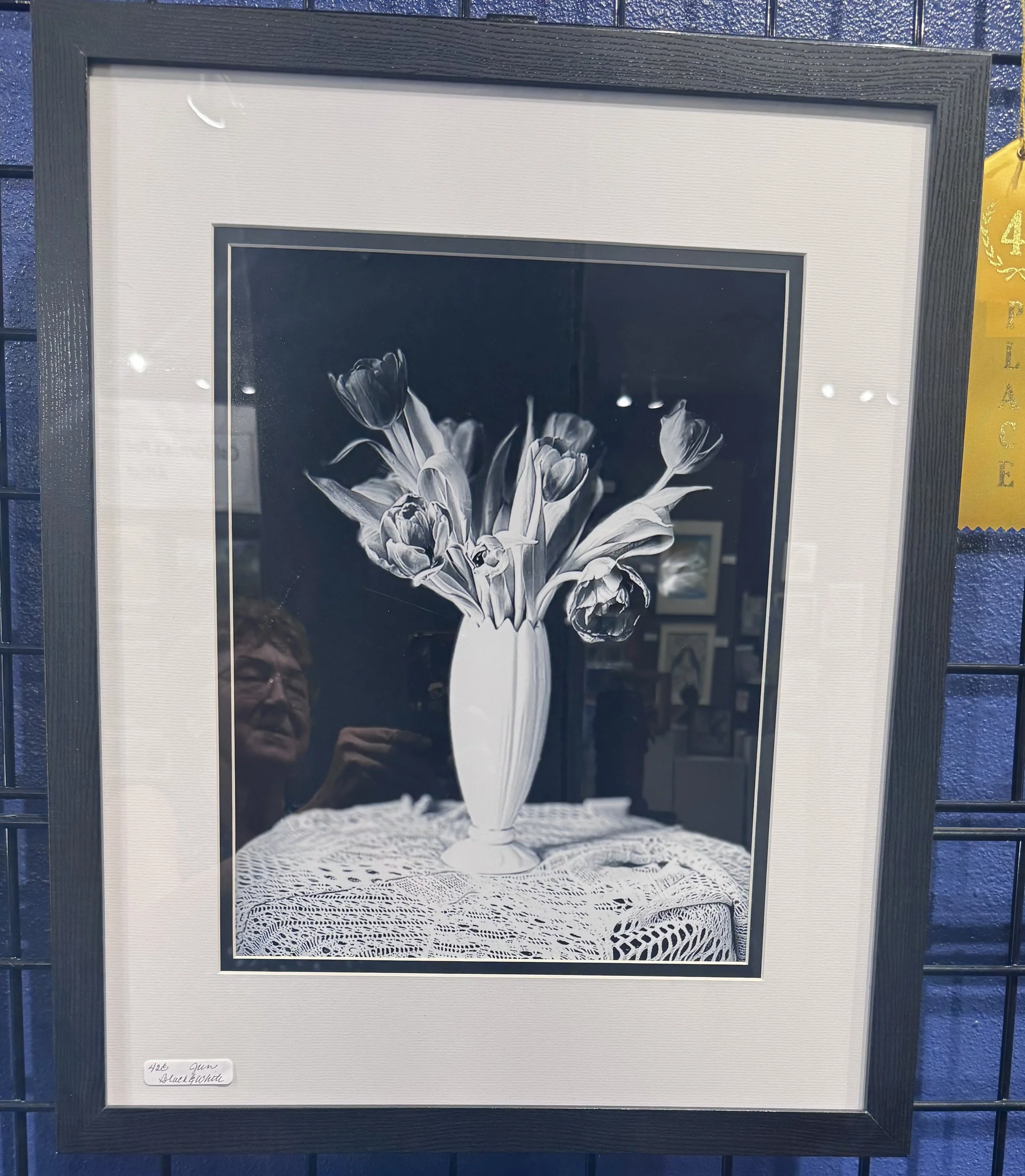 Black-and-white photograph of a vase with tulips on a lace tablecloth, displayed in a black frame with a white mat and a handwritten label that reads '426 Gun Black & White'.