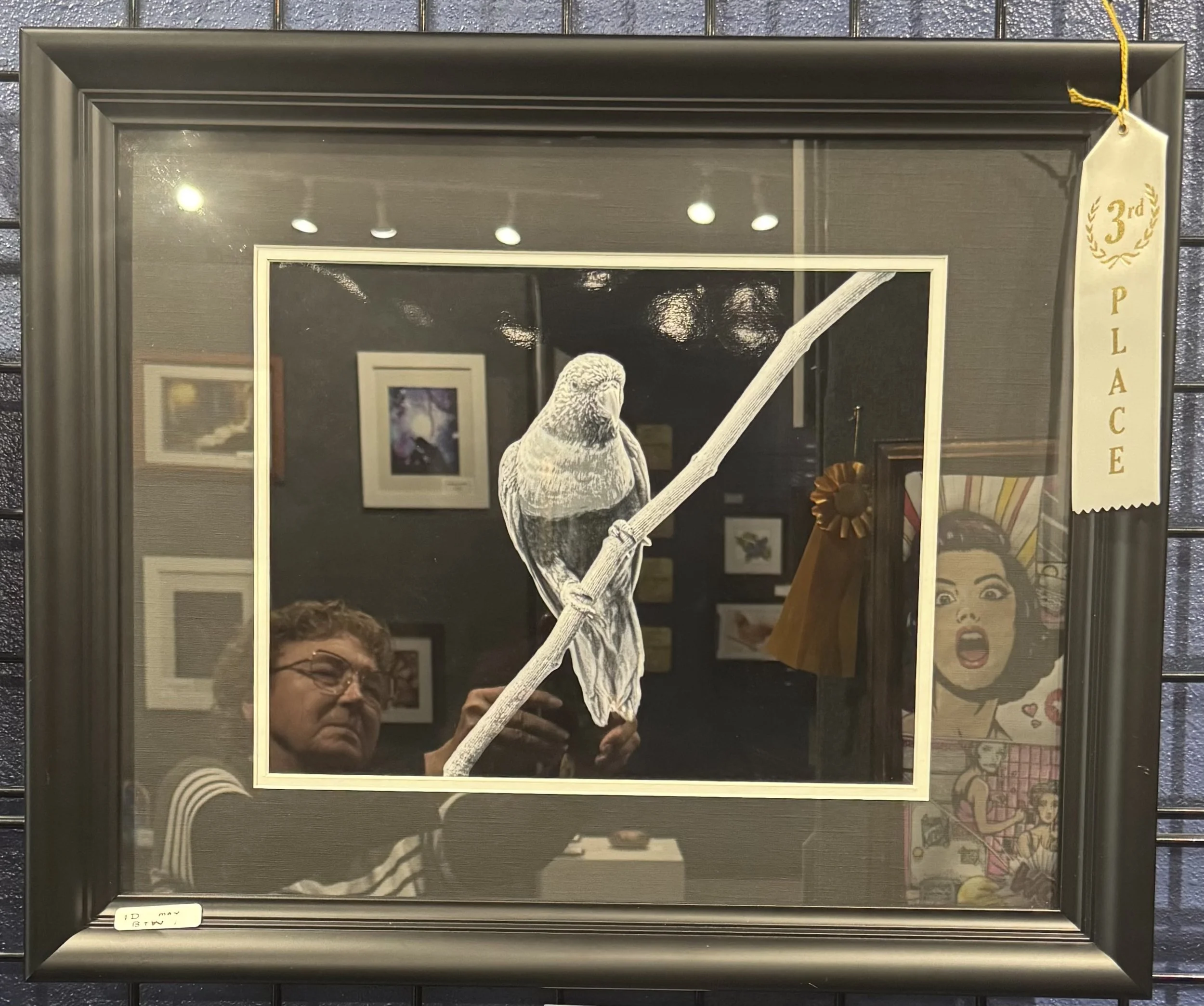 A framed mirror with a sticker on the side that reads "3rd Place". The mirror reflects a man wearing glasses taking a photo, an artwork on the wall, and ceiling lights.