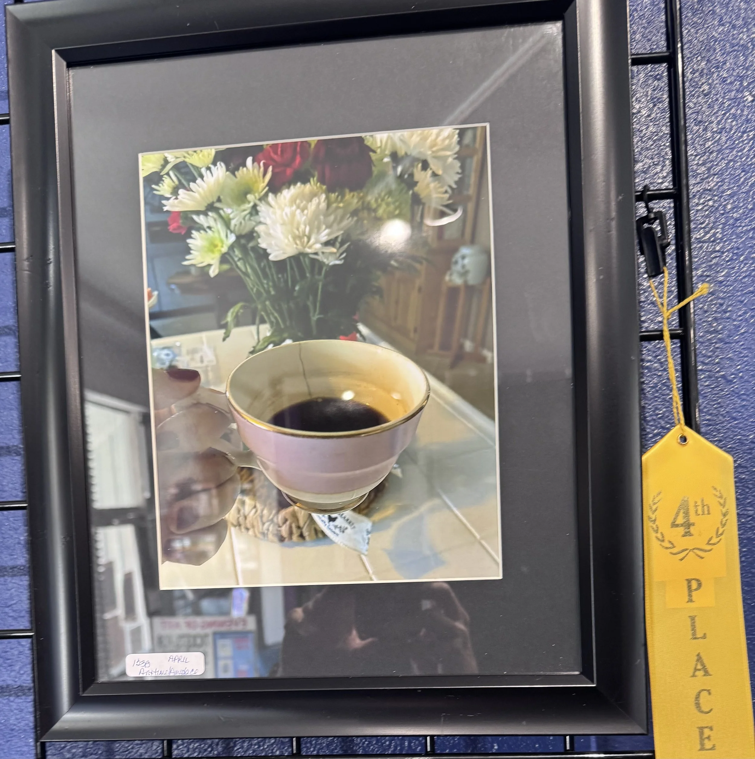 A framed picture of a bouquet of flowers, including white and red flowers and greenery, on a surface with a reflection. Part of a yellow poster and a black wire rack are visible nearby.