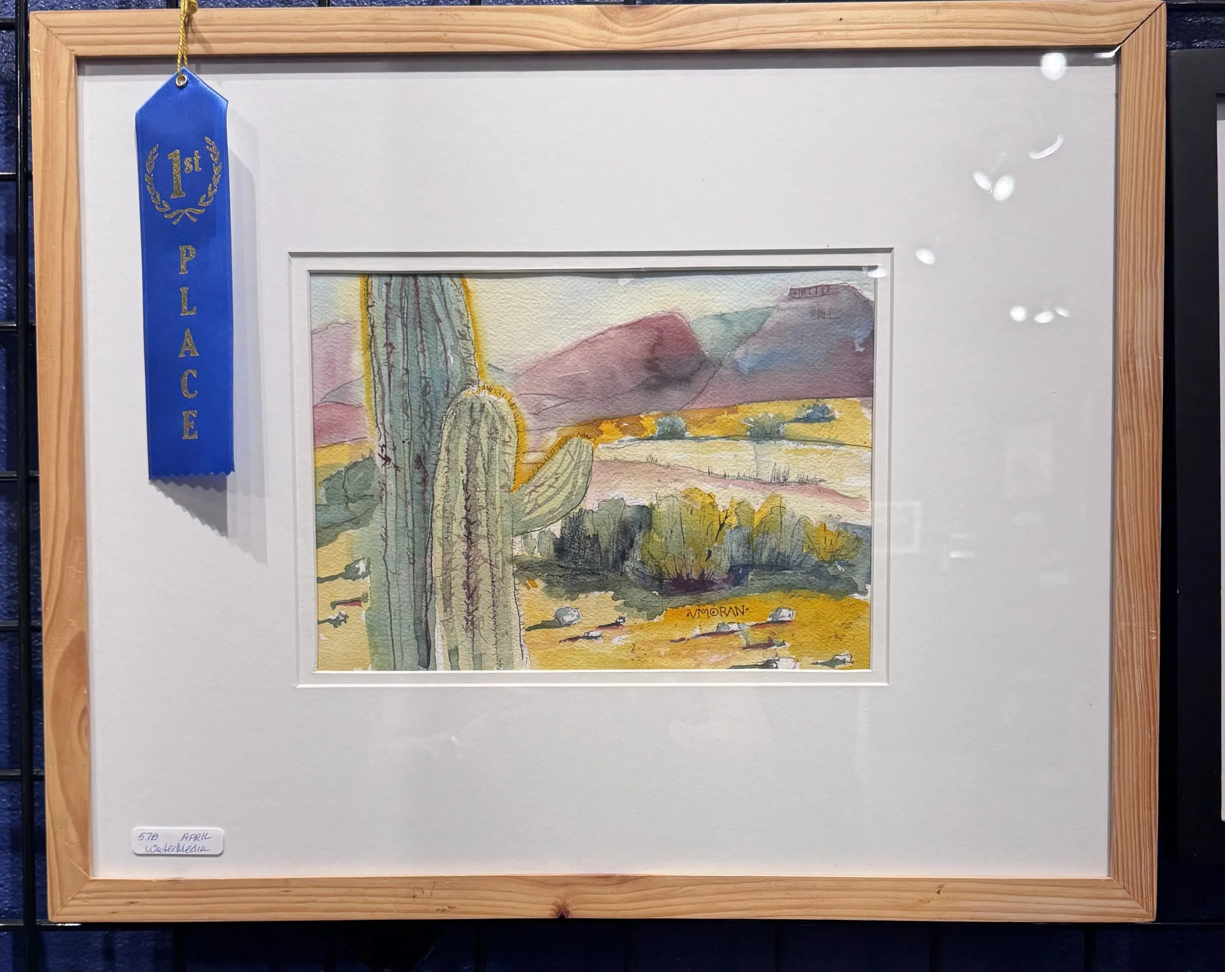 Watercolor painting of desert landscape with cacti, mountains, and a yellow-orange sky, framed with a wooden border and labeled 'April WaterMedia'.