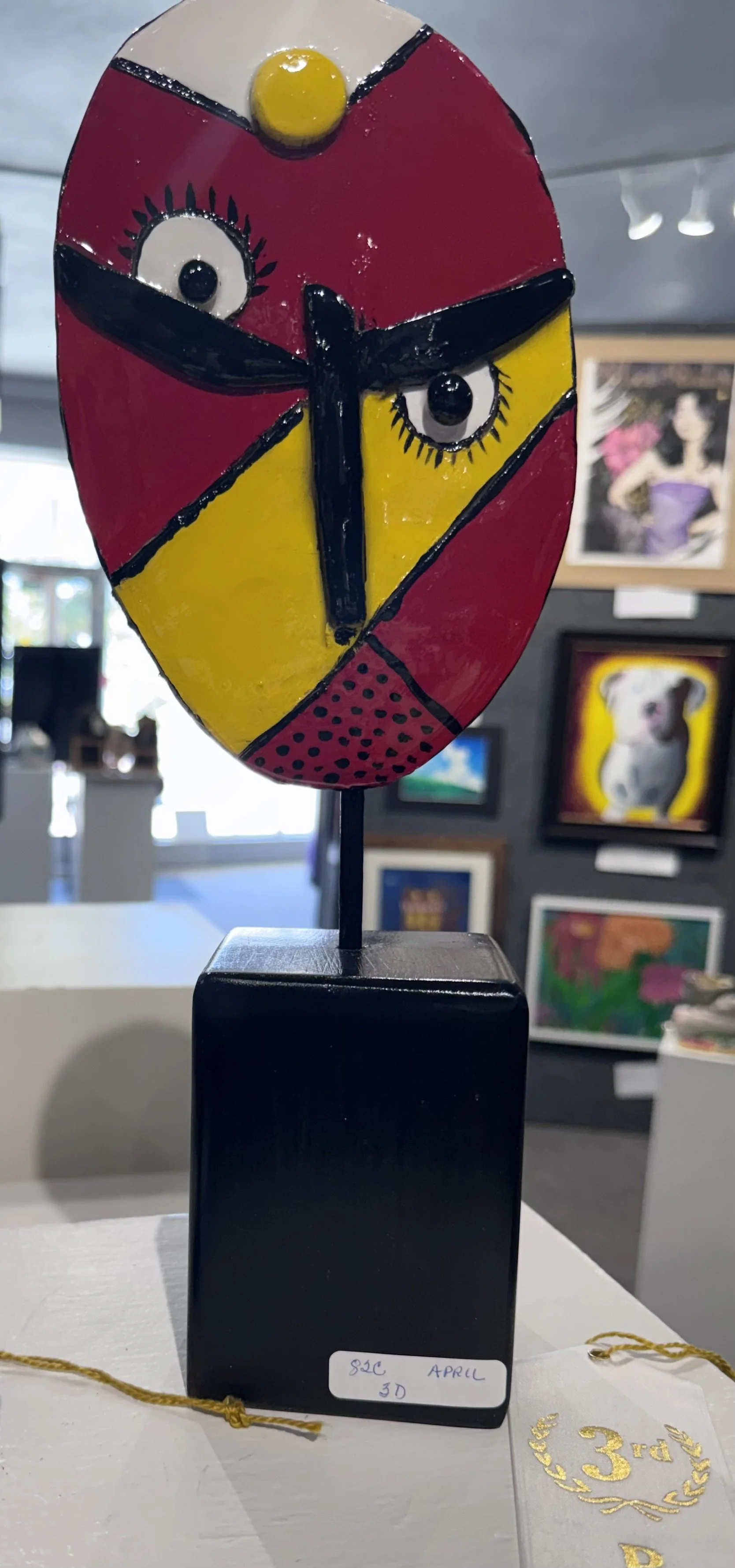 Colorful 3D art sculpture of a face with large eyes, painted in red, yellow, black, and white, displayed on a black pedestal in an art gallery.