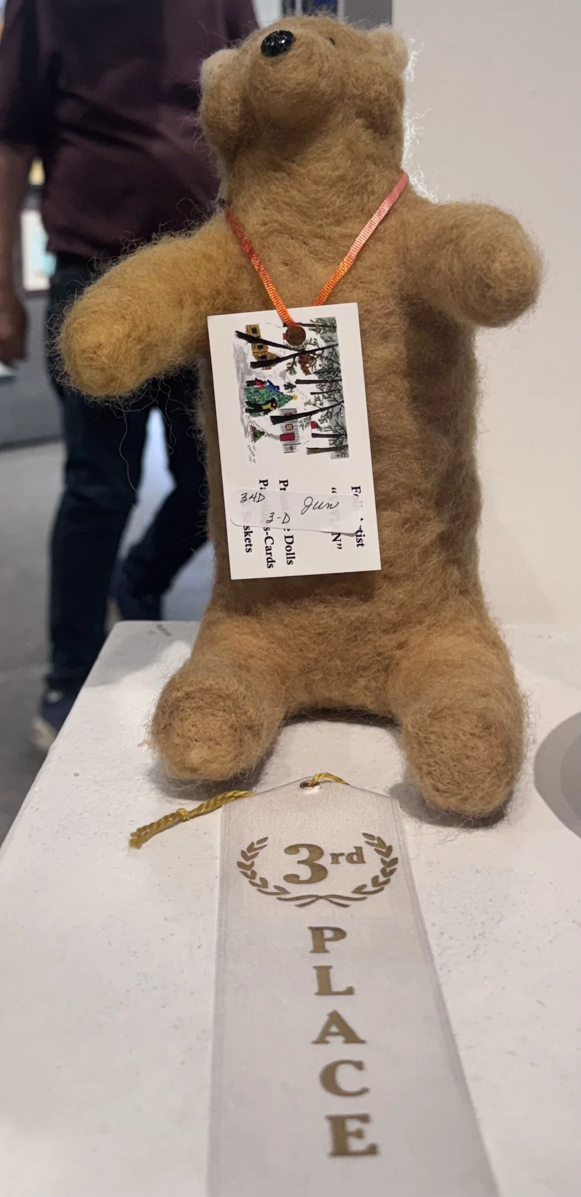 A fuzzy brown teddy bear with a ribbon around its neck, standing on a table with a third-place ribbon sign in front. The bear has a tag hanging from its neck indicating it placed third in a competition.