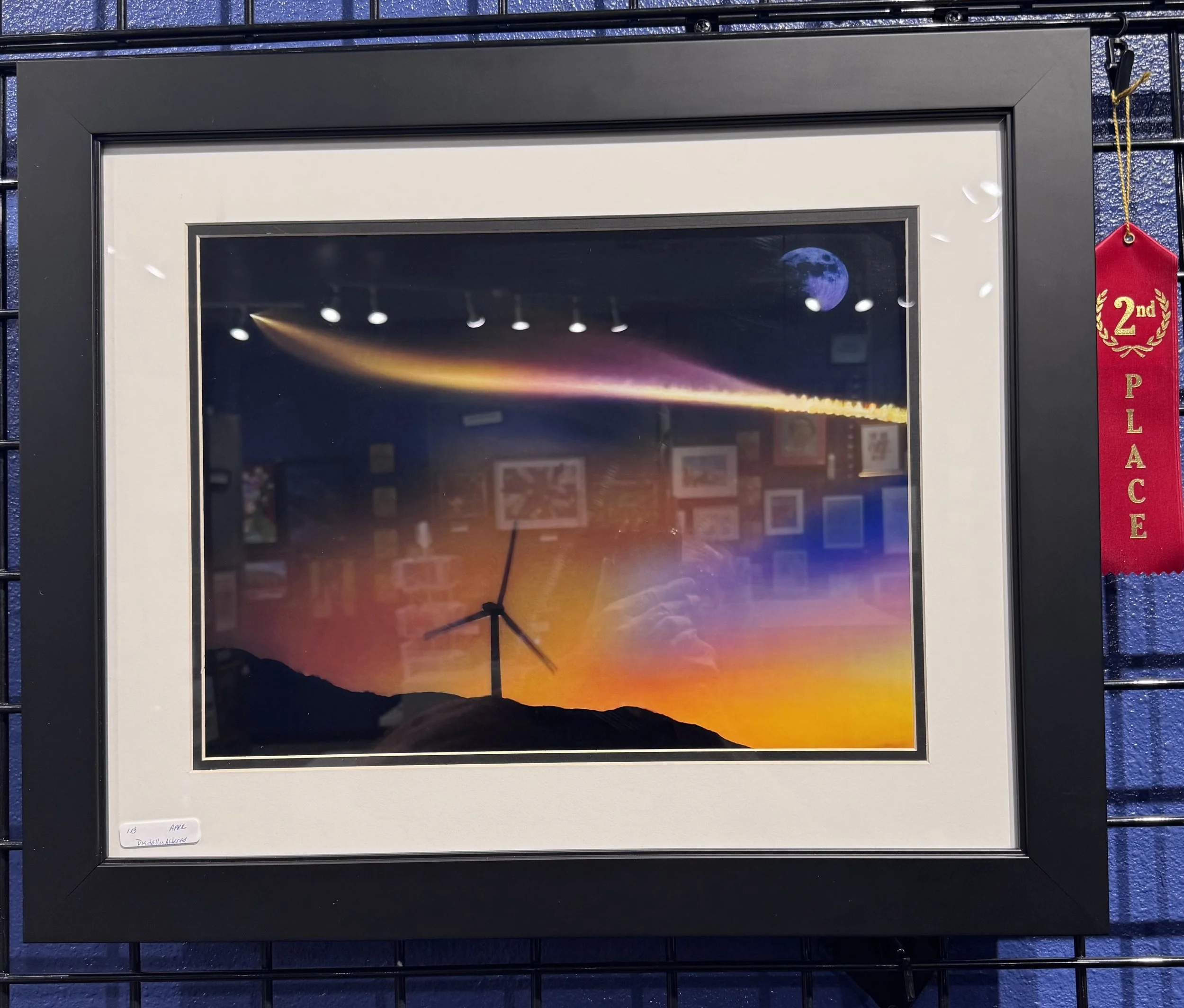 Framed artwork of a sunset with silhouette mountain, wind turbine, and sky with moon and comet, reflected in glass
