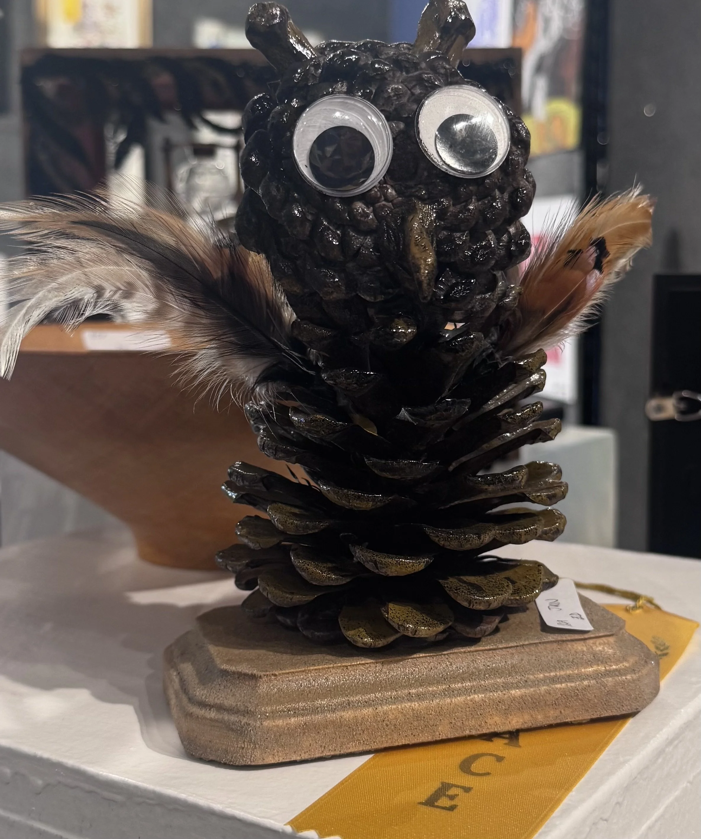 A sculpture resembling a cartoonish owl made of pine cones, with googly eyes and feathers, placed on a round pedestal at a craft show or display.