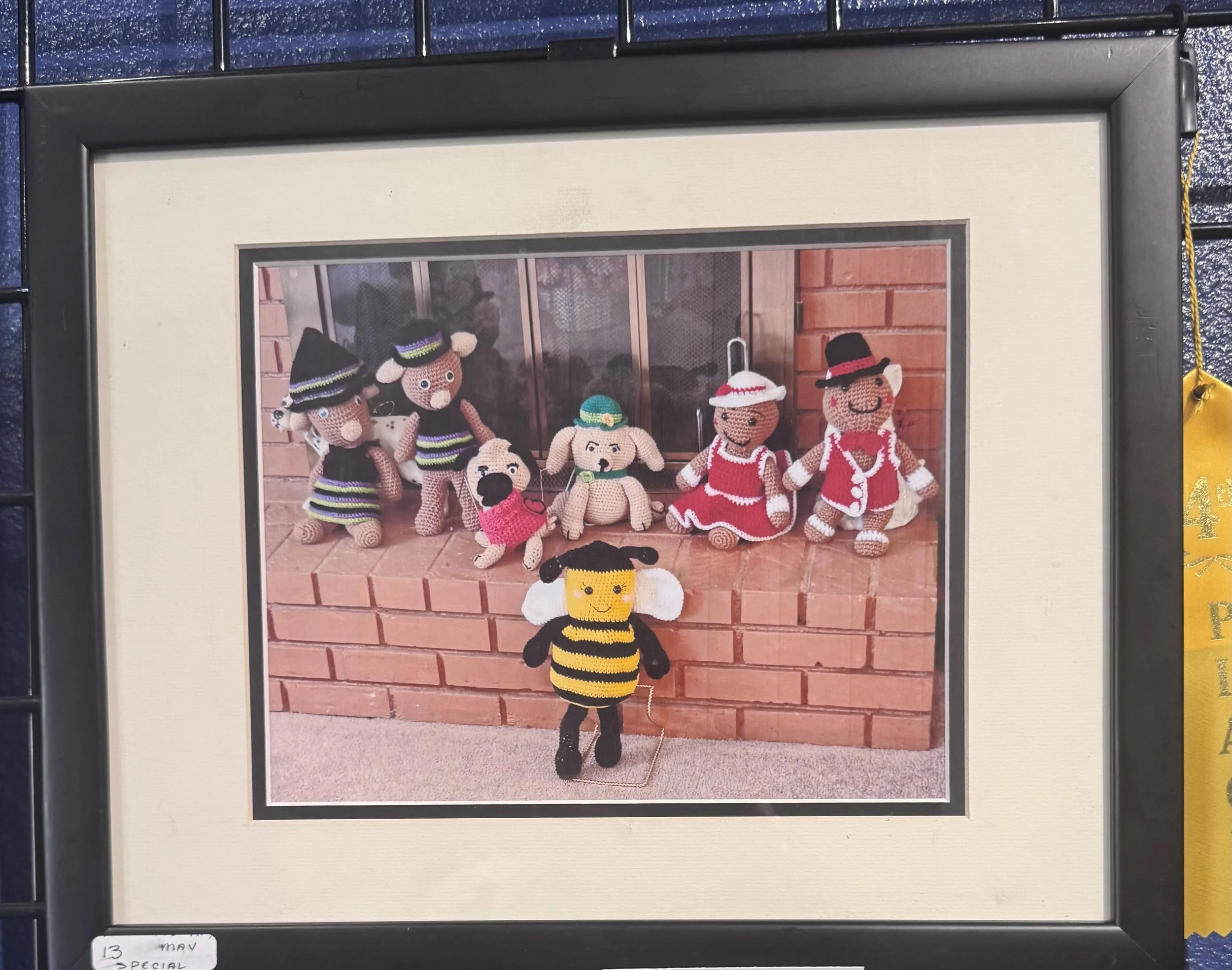 Photograph of handmade crocheted animal dolls dressed in colorful clothes, displayed on a photo board with a brick background.