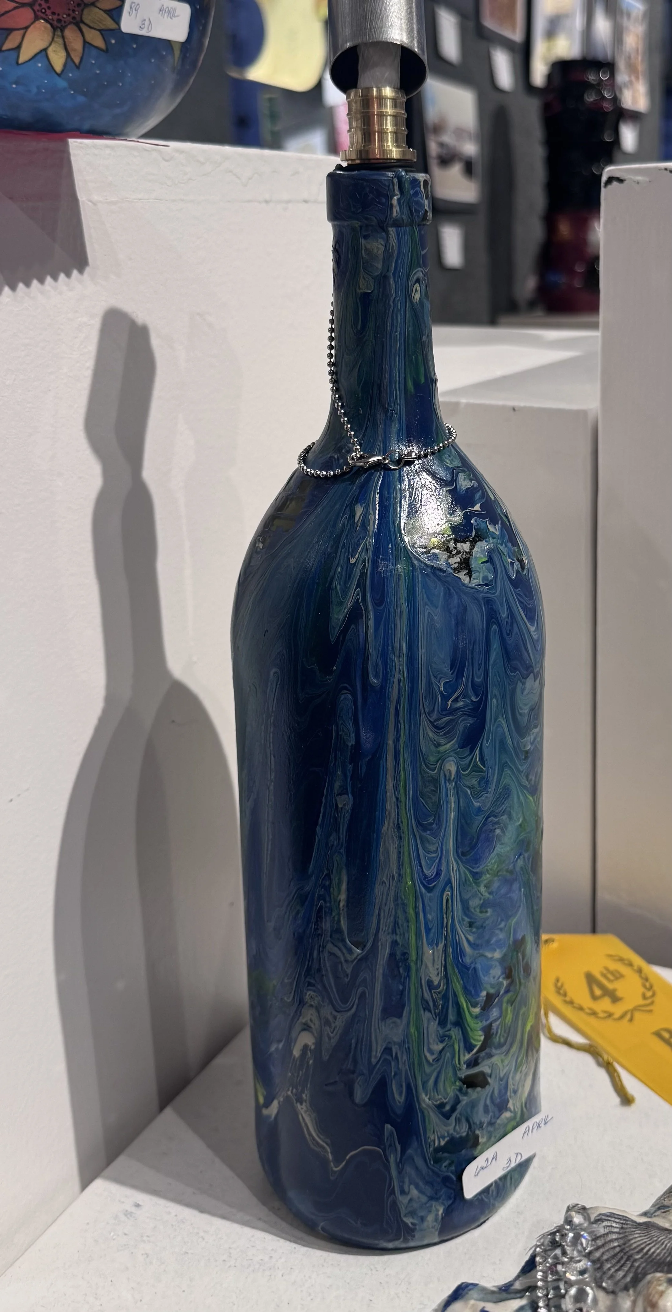 Decorative blue, green, and white marbled glass bottle with a metal cap and chain embellishment on the neck, displayed on a white shelf.