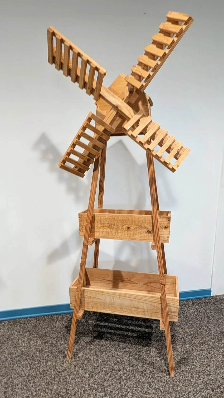 A wooden model of a windmill with four blades, mounted on a stand with two shelves, placed against a plain wall.