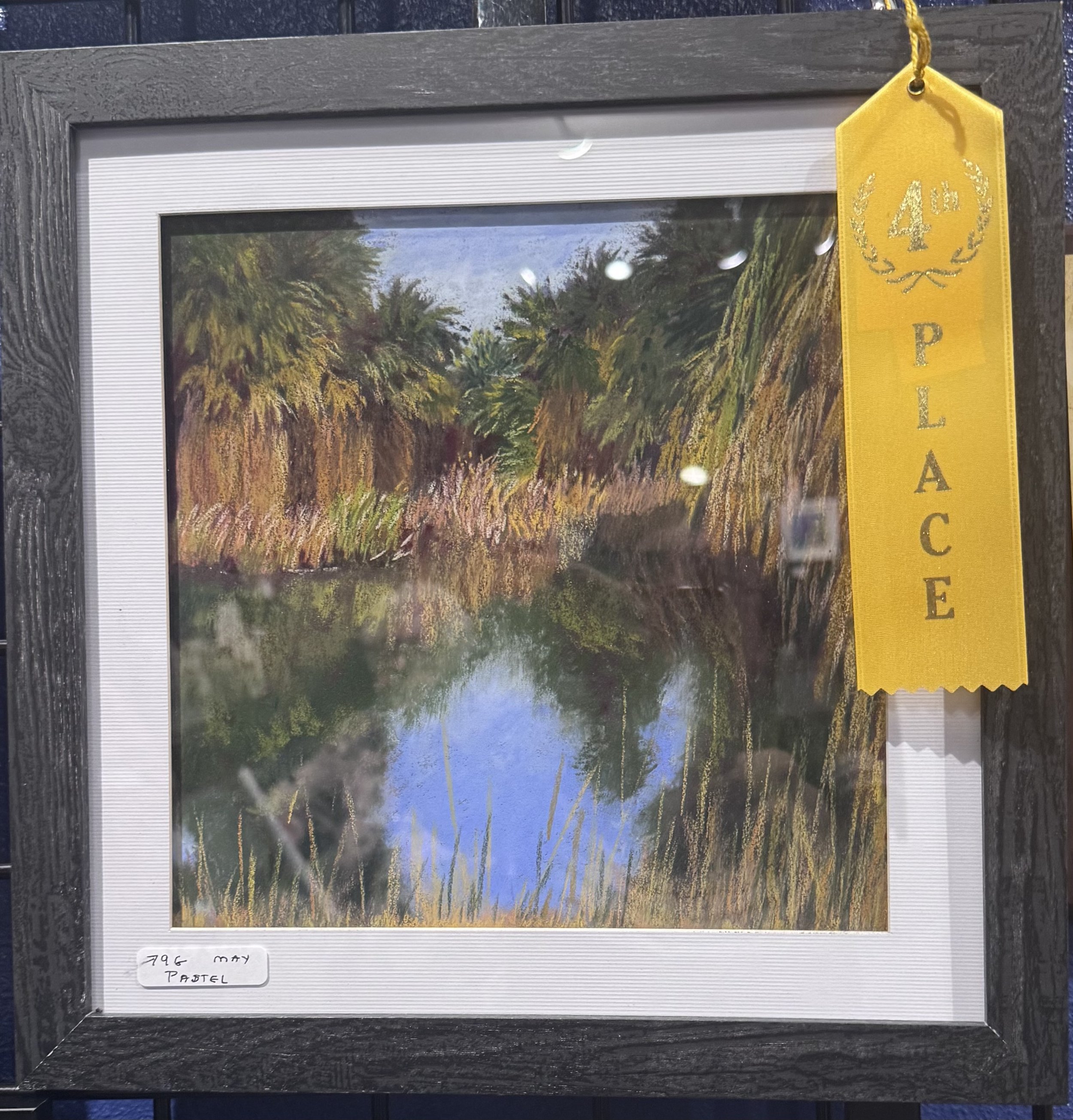 Framed landscape painting of palm trees and greenery with a yellow ribbon on the top right corner labeled with the number 4 and the letter P