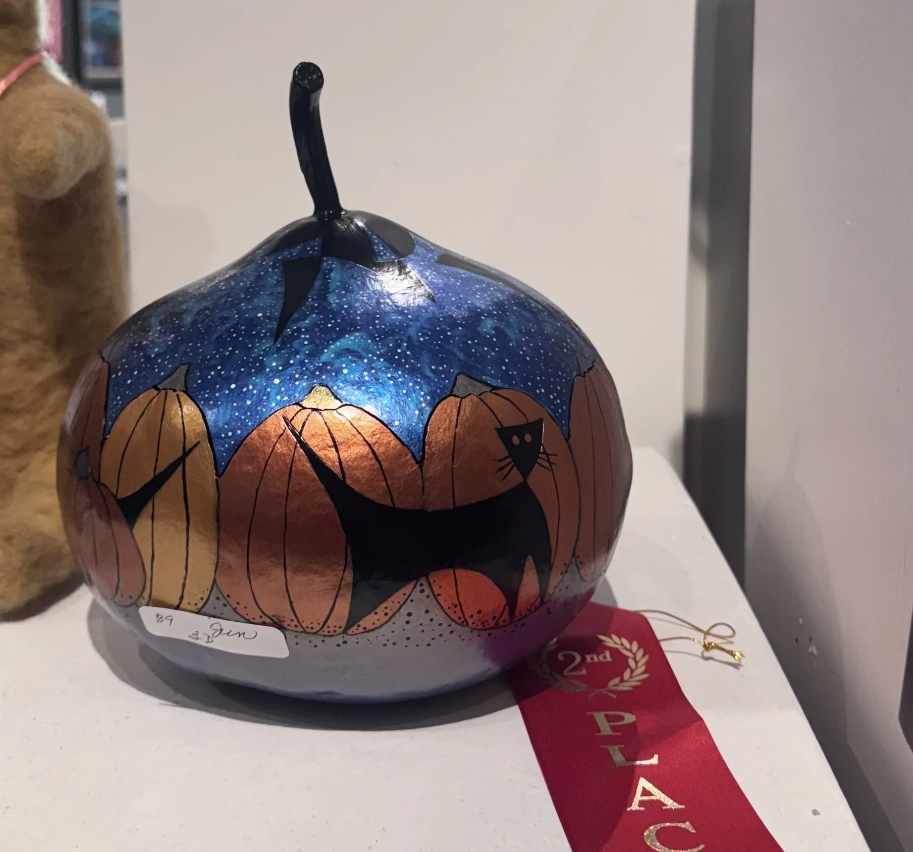A decorative pumpkin-shaped lantern with painted pumpkins and a starry night sky design. It has a black stem and is placed on a white surface, partly revealing a red ribbon with gold lettering and a gold string accessory.