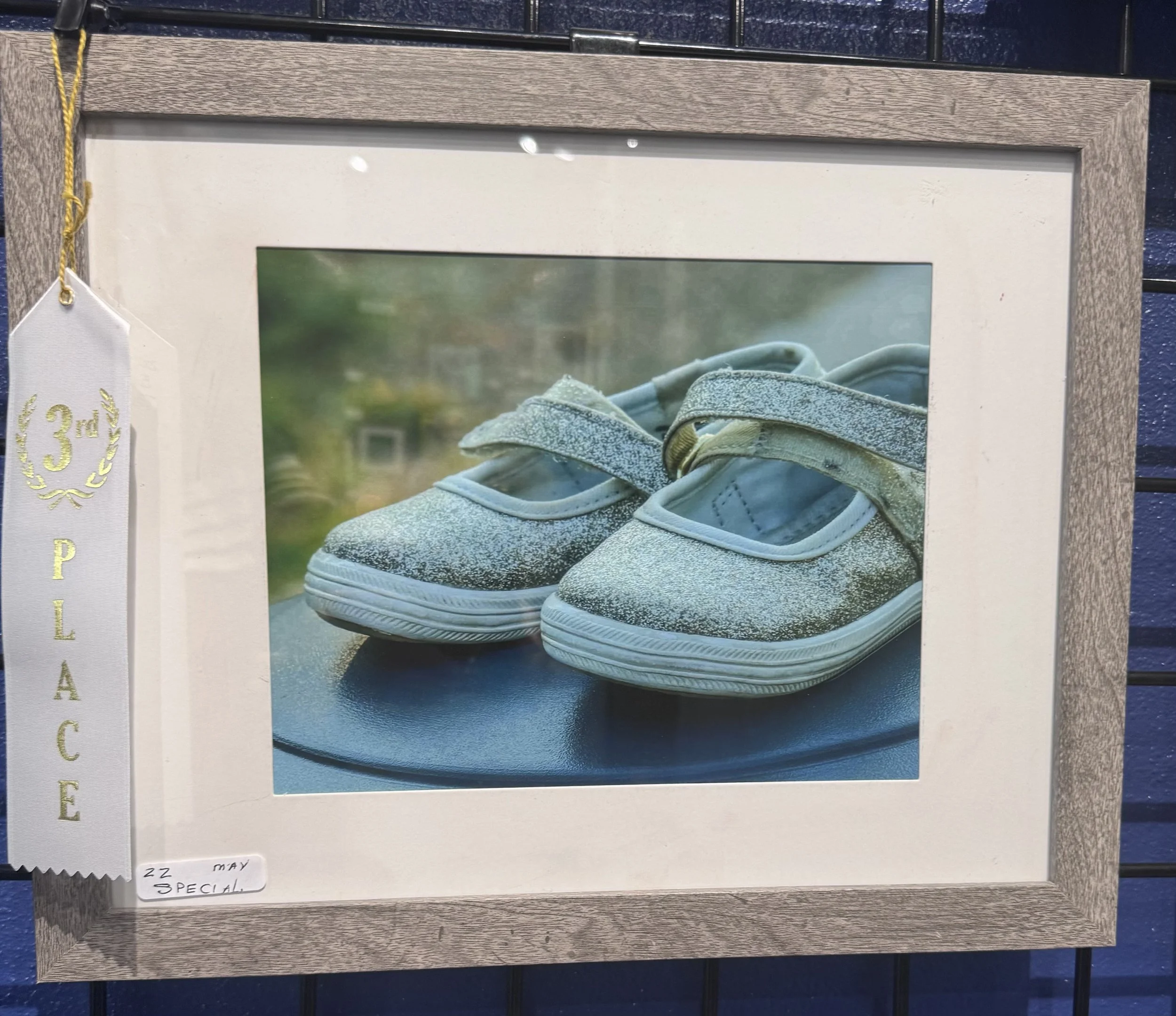 Photo of a framed picture of blue sneakers with a white tag indicating third place in a competition, on a wooden surface with a dark blue tiled background.
