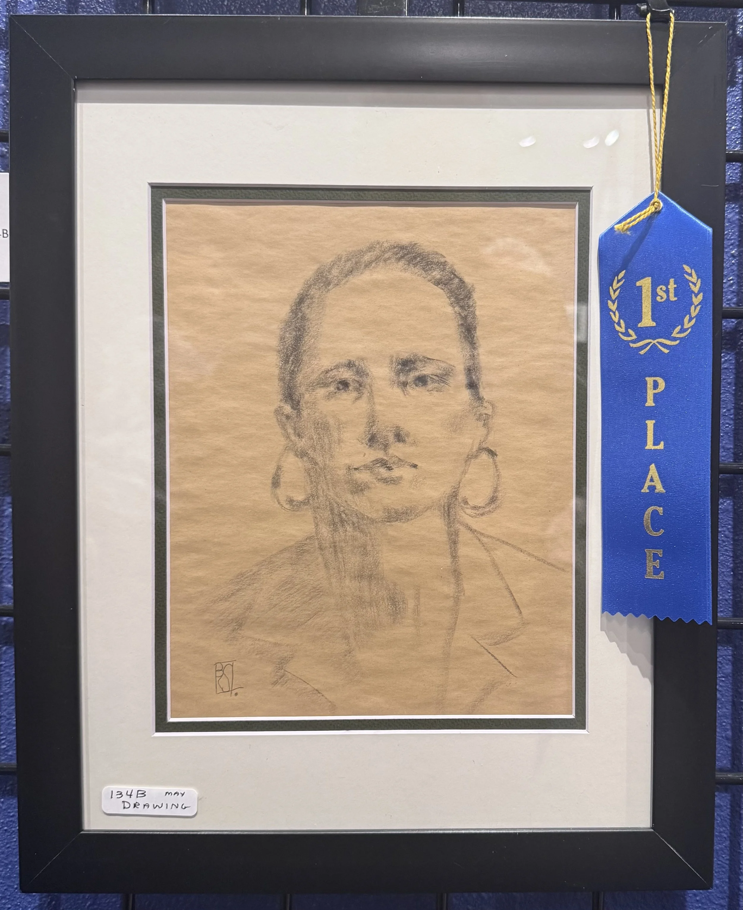 Framed pencil sketch of a woman's face with earrings, titled '134B May' with a blue first place ribbon hanging on the upper right corner.