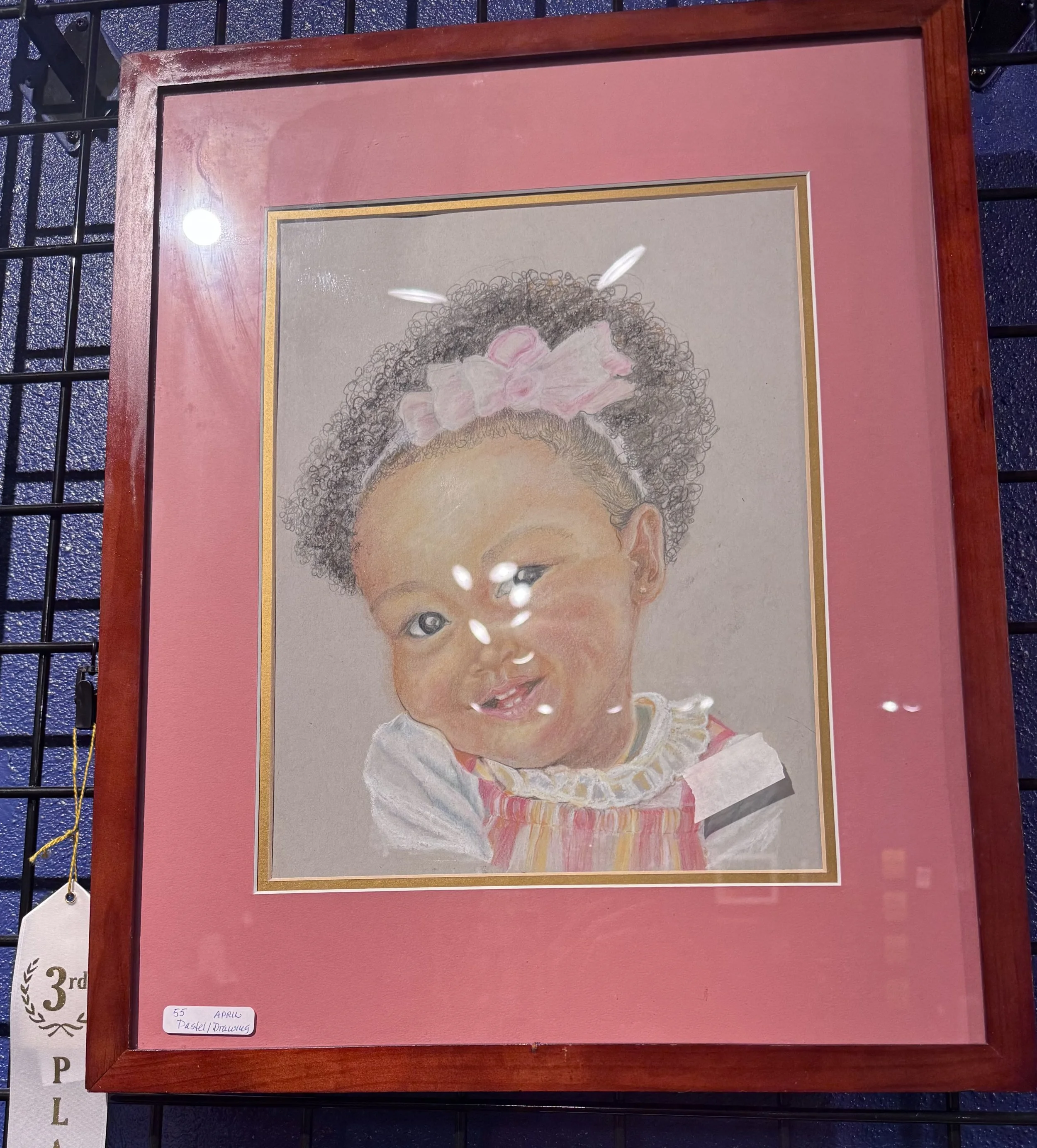 A framed pastel drawing of a young girl with curly hair, a pink bow, and a colorful dress, displayed on a pink background at an art competition.