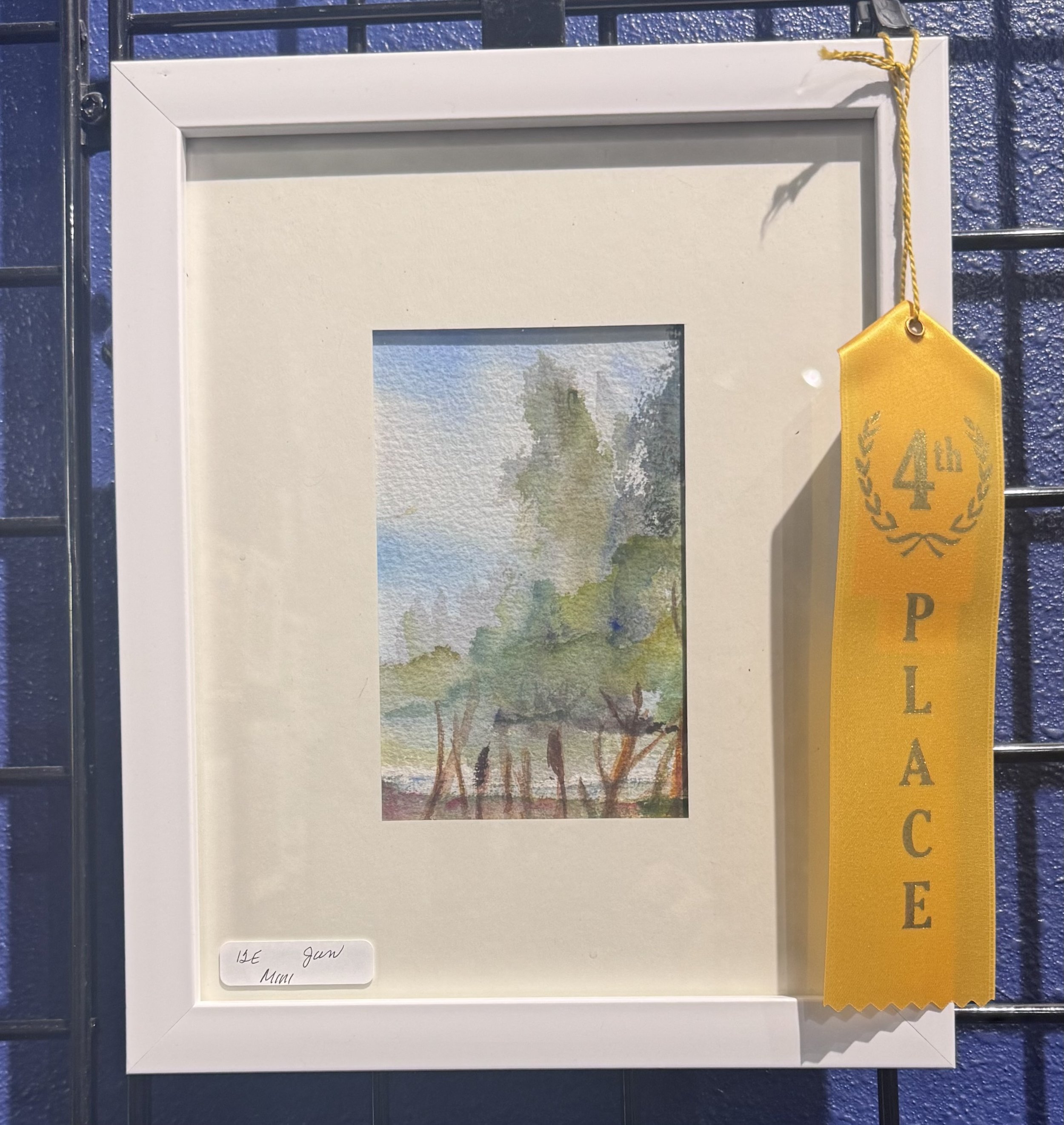 Artwork of trees with green leaves on a watercolor painting, framed in white, displayed on a blue wall with a yellow ribbon attached.