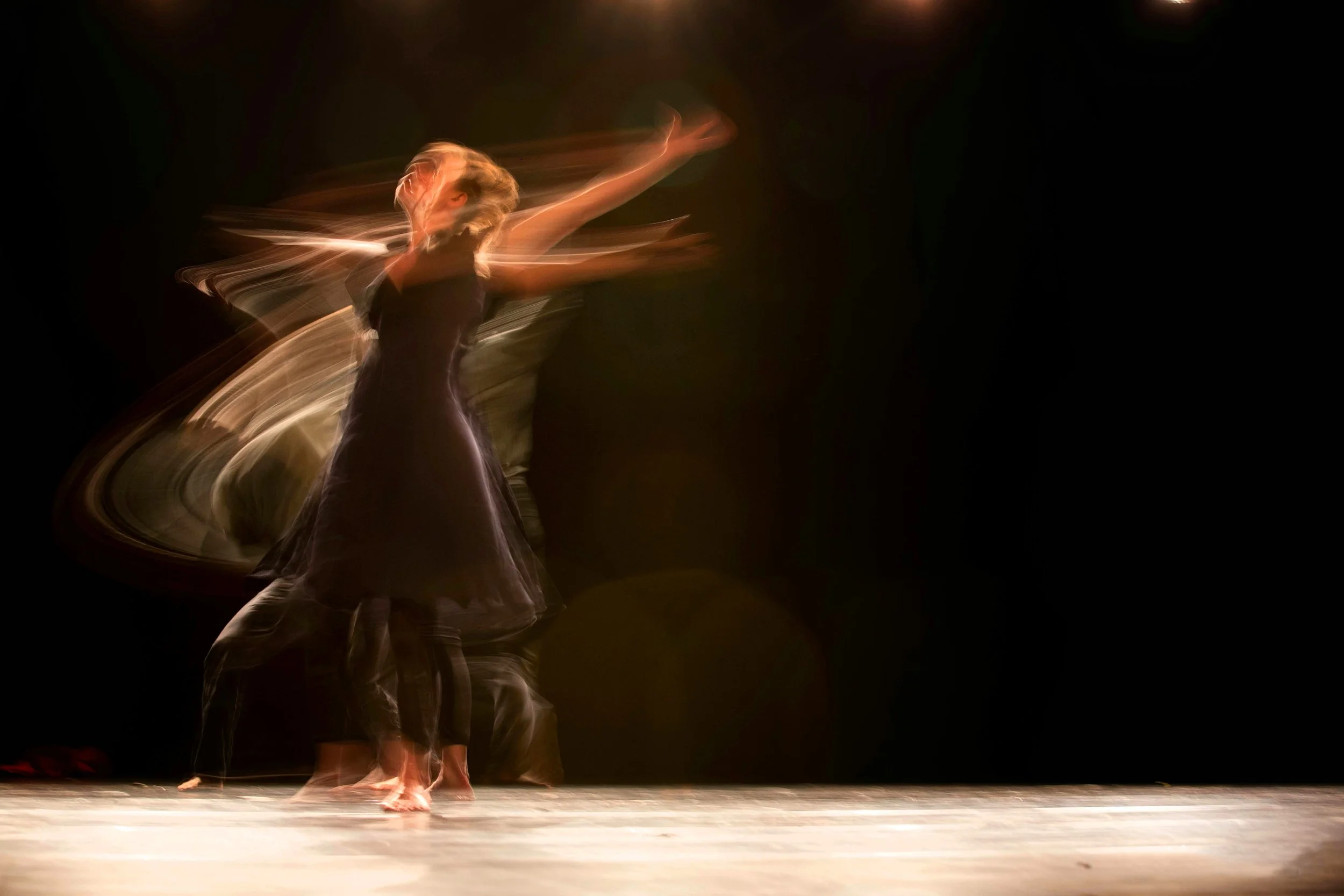 A ballerina dancing on stage with motion blur, wearing a black dress and ballet slippers, captured mid-movement against a dark background.