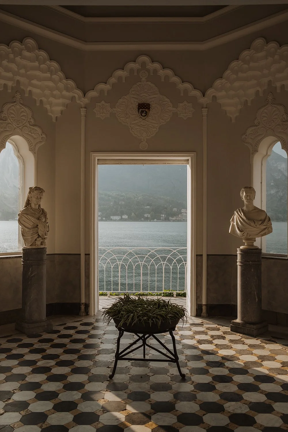 An elegant room with intricate white architectural details, two marble bust sculptures on pedestals, a small table with a potted plant, and an open doorway providing a view of a lake and mountains in the background.