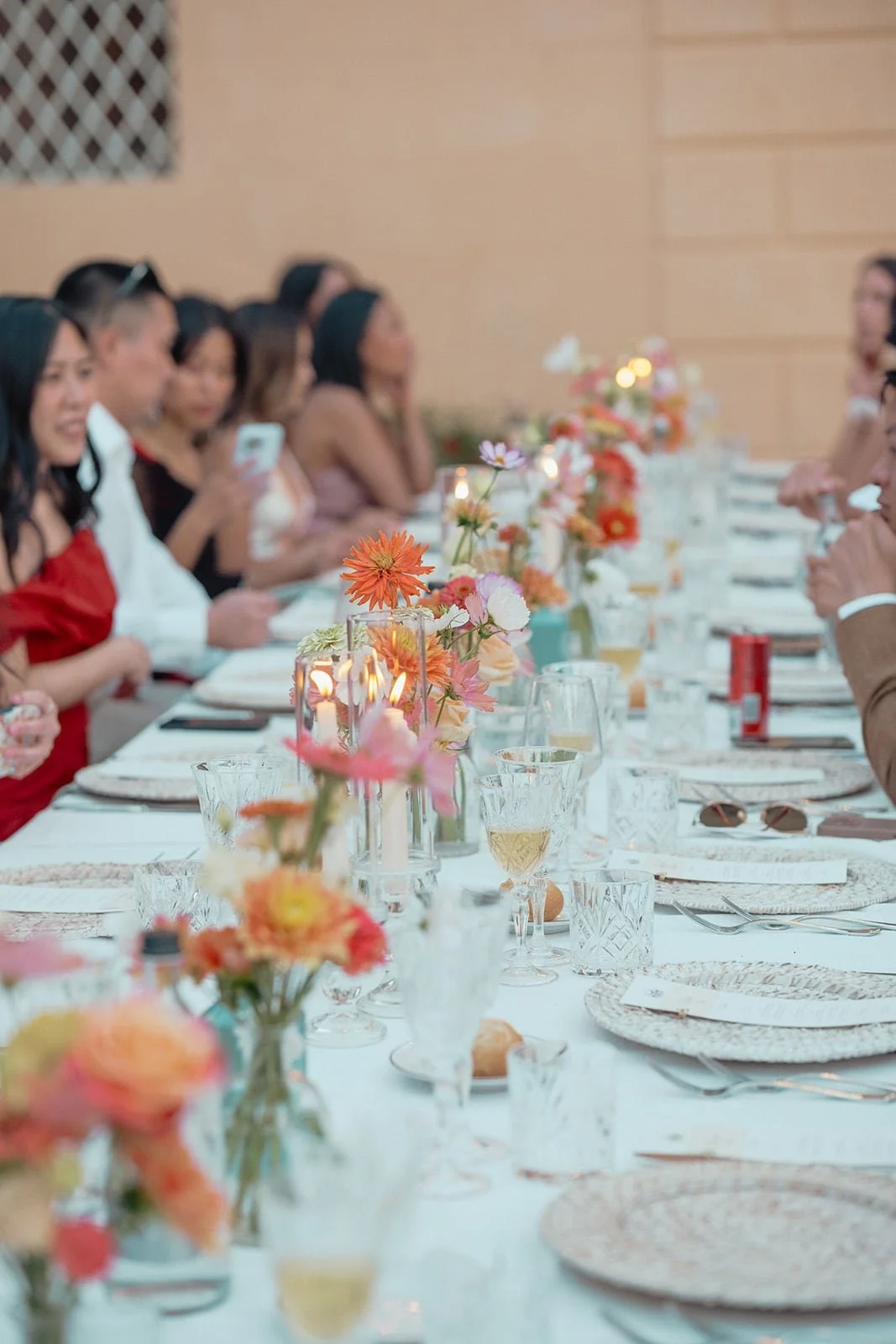 A side-angle shot of the wedding guests seated at the long dining table, focused on the vibrant floral centerpieces and the warm, social atmosphere of the dinner.