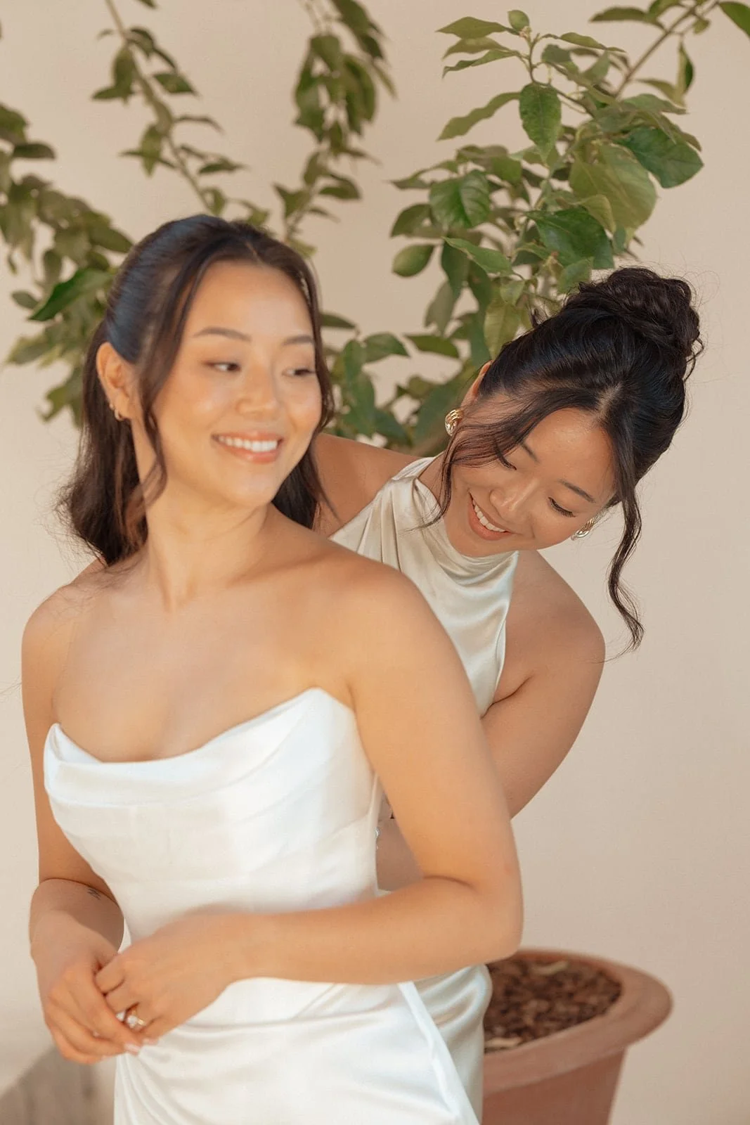 The bride smiles as her bridesmaid gently adjusts the dress, sharing a calm and joyful moment before the ceremony.
