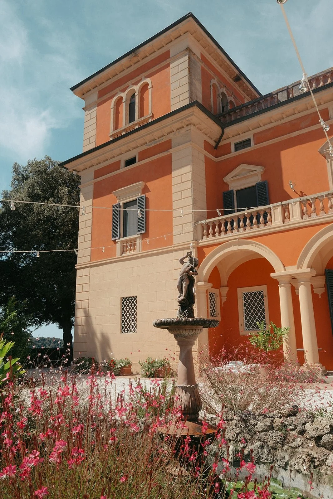 The Villa Lena façade framed by summer blooms and a stone fountain, photographed in early September in Tuscany.