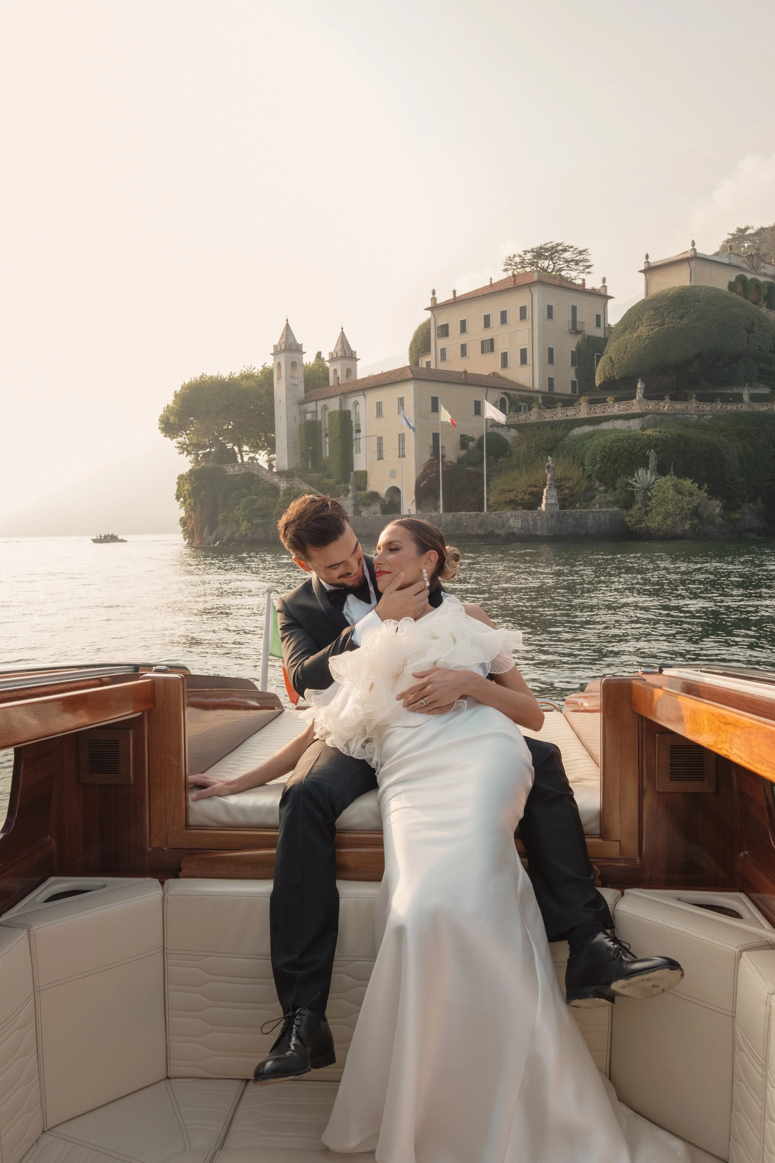 A bride and groom embracing on a wooden boat at Lake Como, with an elegant Italian villa in the background, photographed in a refined editorial wedding style.