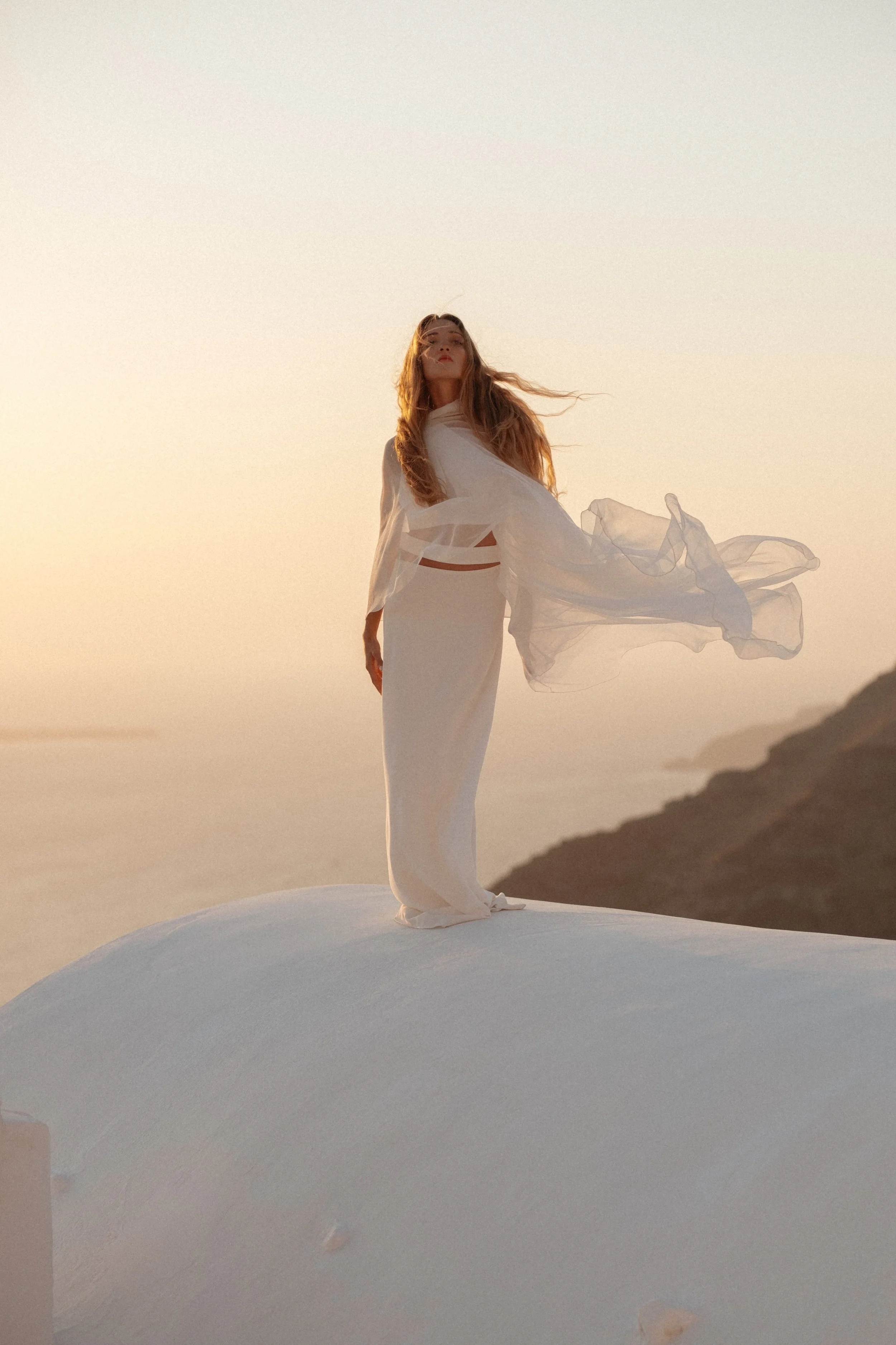 An editorial bridal portrait in Greece, capturing modern elegance, soft movement, and cinematic light overlooking the Mediterranean landscape.