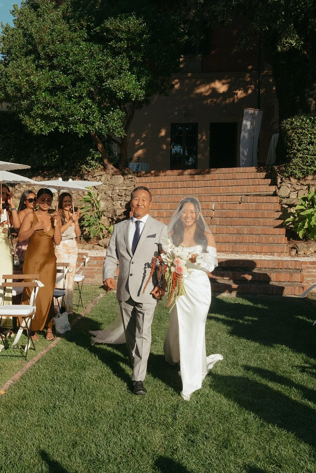 The bride and her father make their entrance together, surrounded by guests and warm afternoon light at Villa Lena.