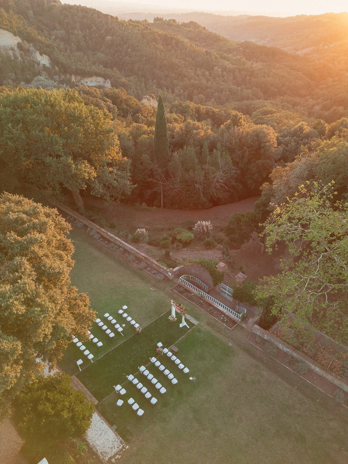 An aerial view of the ceremony setup at Villa Lena, surrounded by rolling Tuscan hills and bathed in golden sunset light.