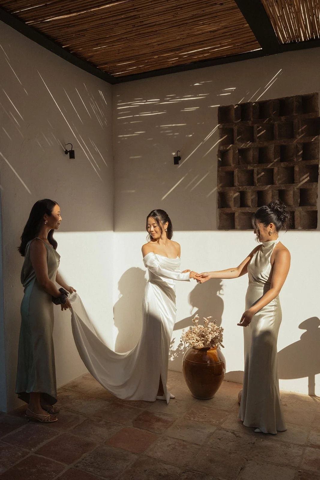 Sunlight streams across the room as the bride stands with her bridesmaids, sharing an intimate moment before the wedding.
