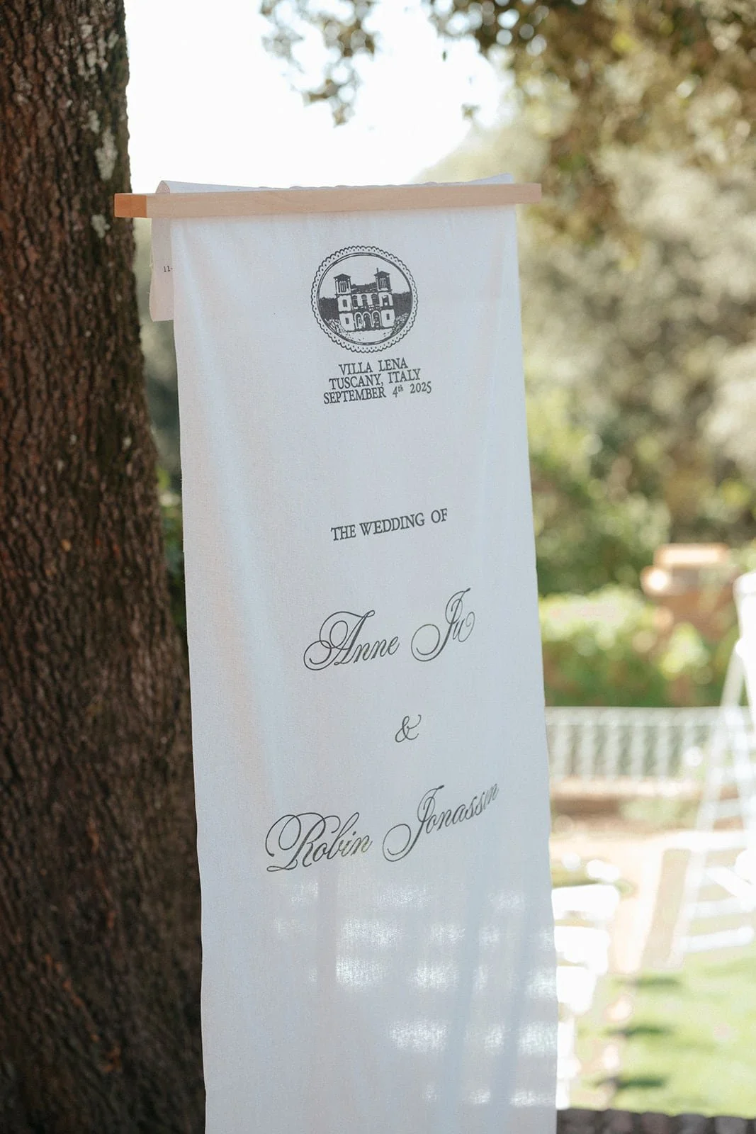 A linen wedding banner displaying the couple’s names and date, hanging beneath the trees at Villa Lena in the Tuscan countryside.