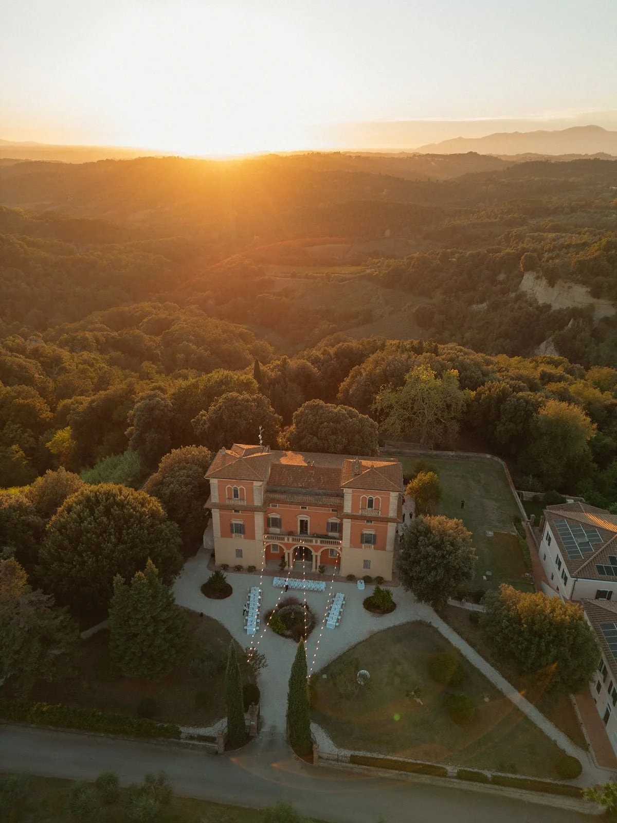 An aerial view of the Villa Lena estate during the golden hour, highlighting the outdoor dining area arranged in a U-shape in the courtyard and the surrounding forest.