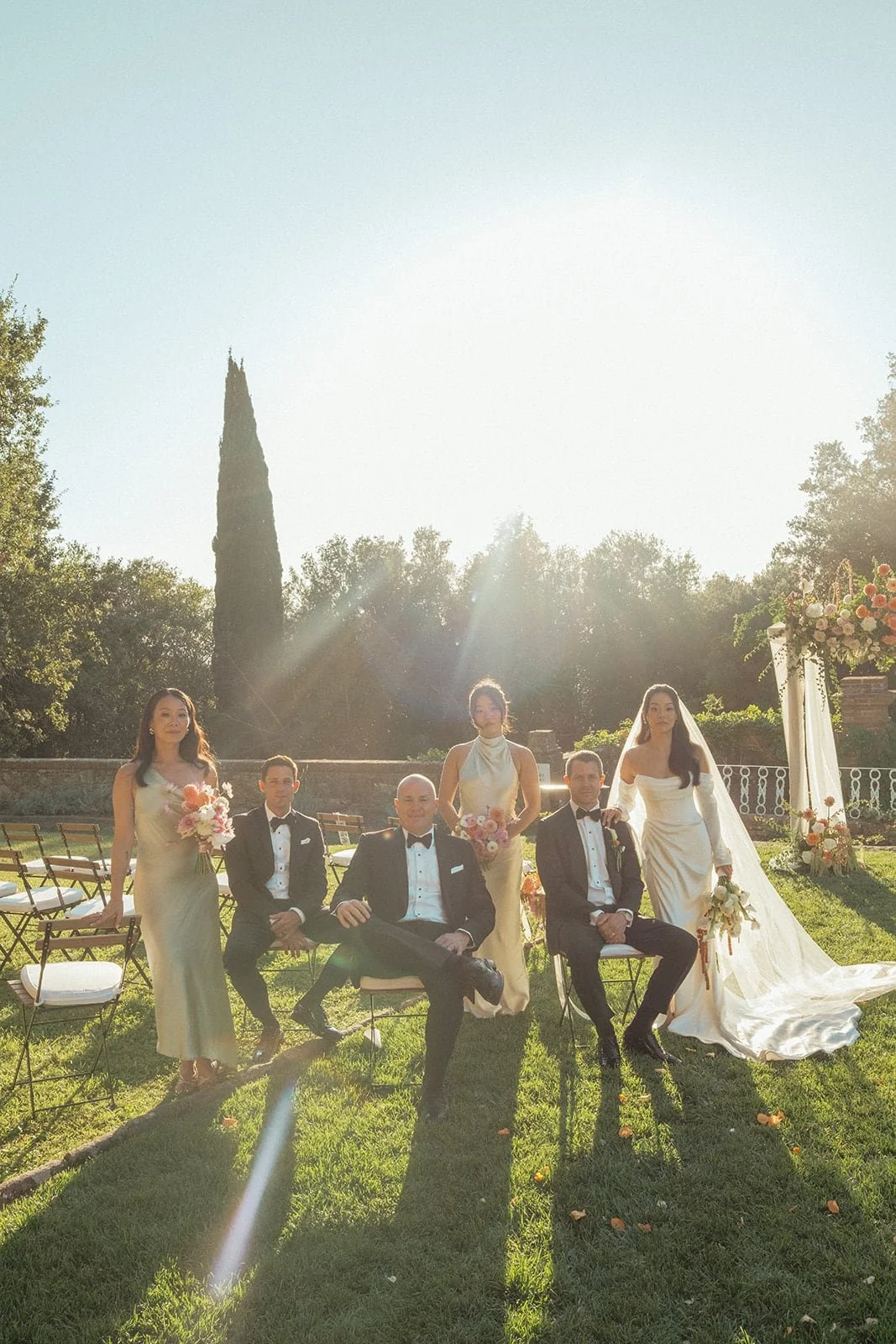A relaxed portrait of the wedding party seated on the lawn, captured in soft summer light with the ceremony setting behind them.