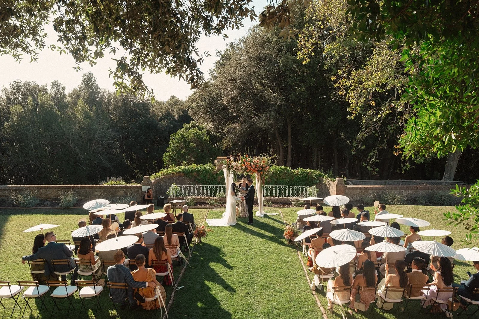 A wide aerial-style view of the outdoor wedding ceremony at Villa Lena, with guests seated under parasols and the couple exchanging vows in the Tuscan landscape.