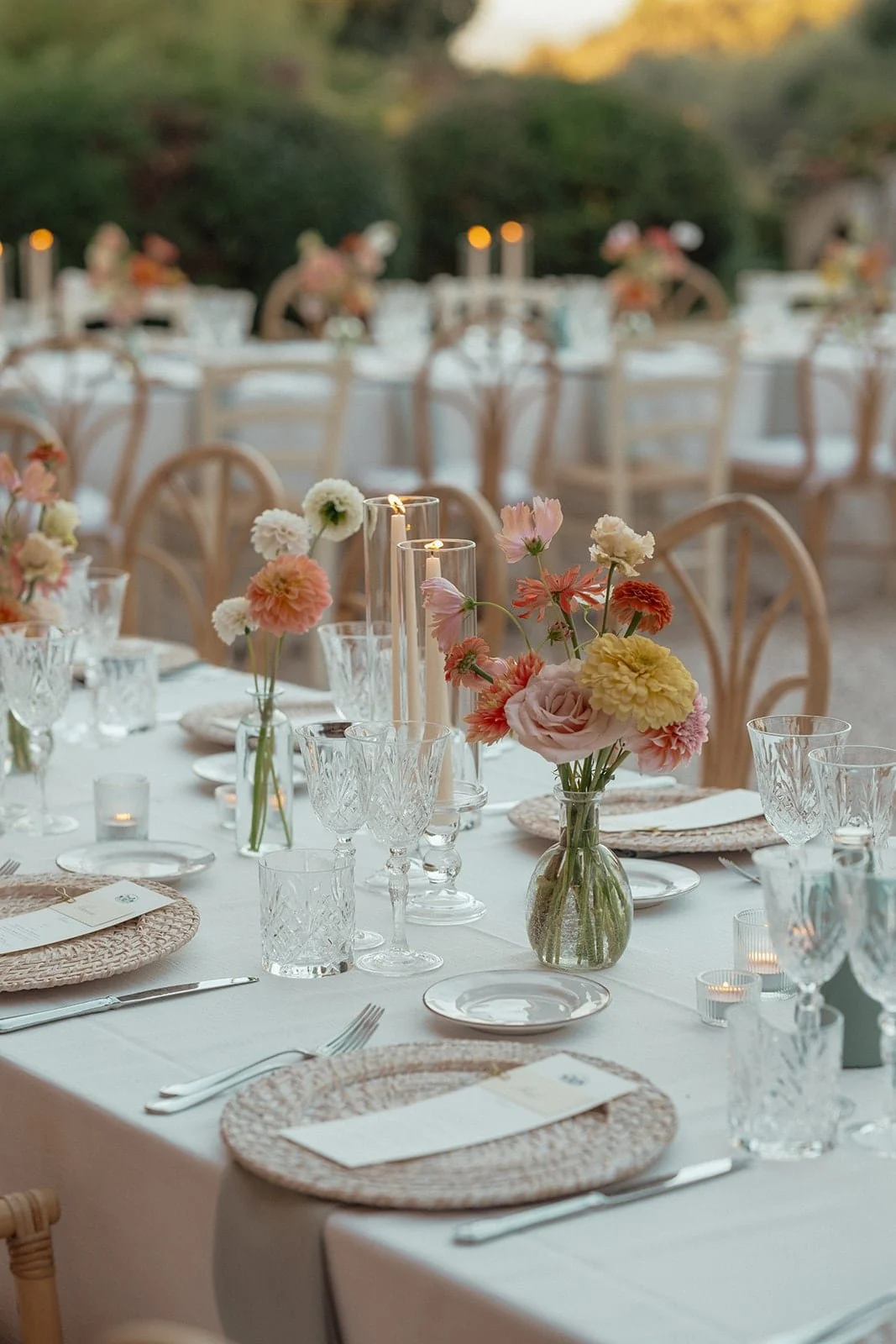 A close-up, shallow-depth-of-field shot of a long wedding reception table featuring elegant crystal glassware, tall taper candles in glass chimneys, and wildflower arrangements in small glass vases.