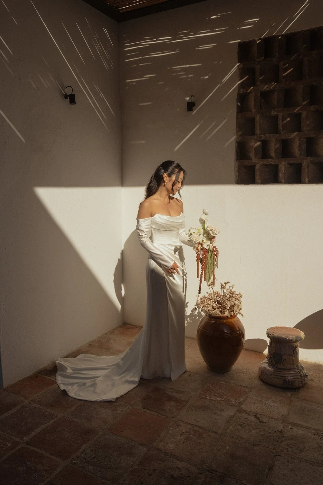 The bride holds her bouquet in soft afternoon light, framed by shadows and textures inside a contemporary villa.