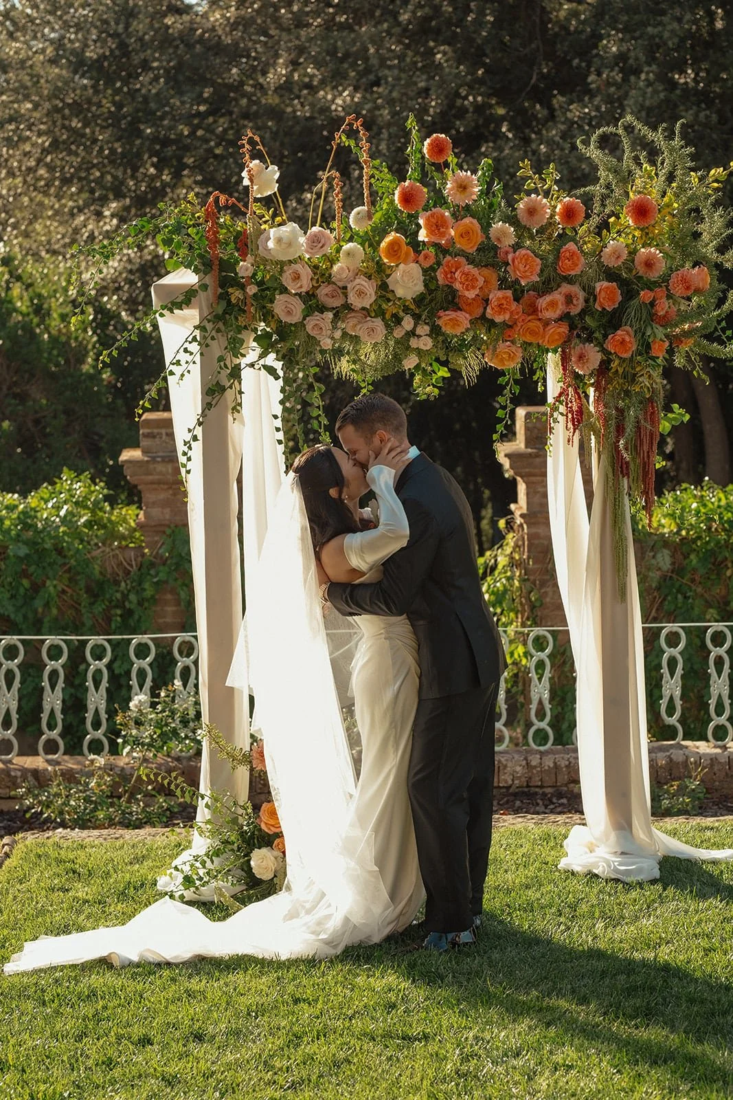 The couple share their first kiss beneath a lush floral arch during their outdoor wedding ceremony in Tuscany.