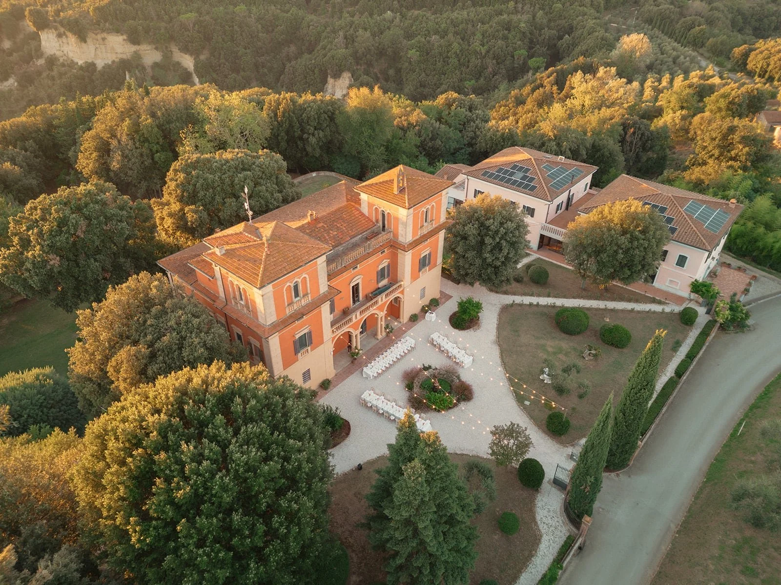 An aerial drone photograph of Villa Lena at sunset, showing the terracotta-colored villa nestled in the lush Tuscan hills with the sun dipping below the horizon.