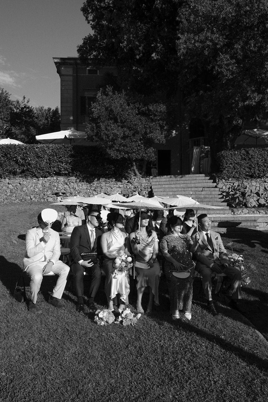 Guests sit together during the ceremony, photographed in black and white with parasols creating a rhythmic pattern across the lawn.