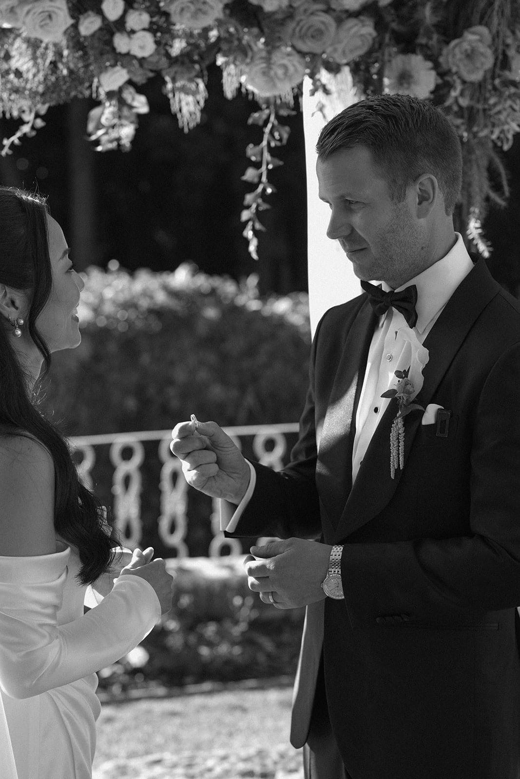 A black and white moment of the groom holding the ring during the ceremony, framed by floral details and soft natural light.
