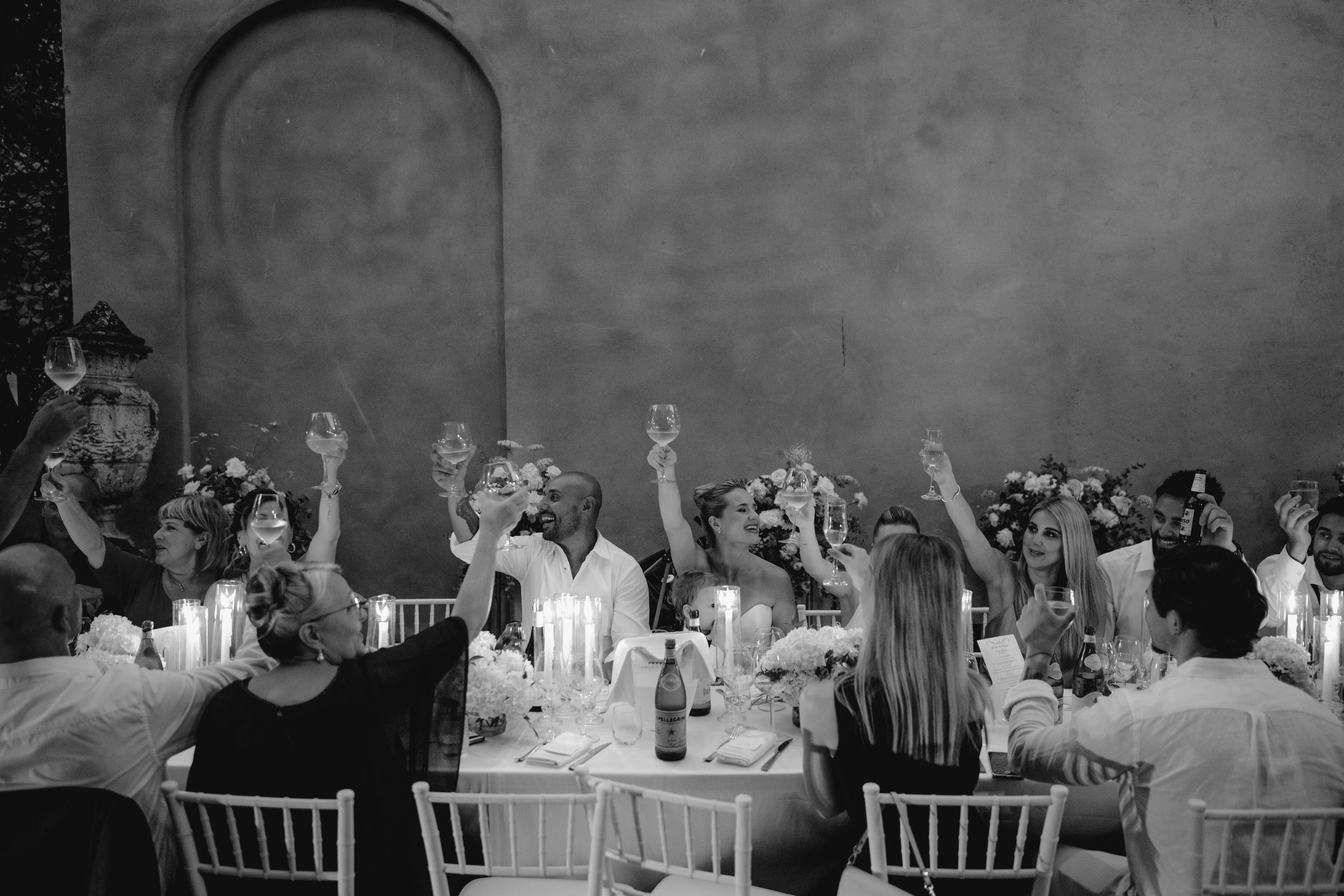 An intimate luxury wedding dinner in Rome, captured in a timeless black and white moment as guests raise a toast during an elegant, candlelit celebration in a historic Italian setting.