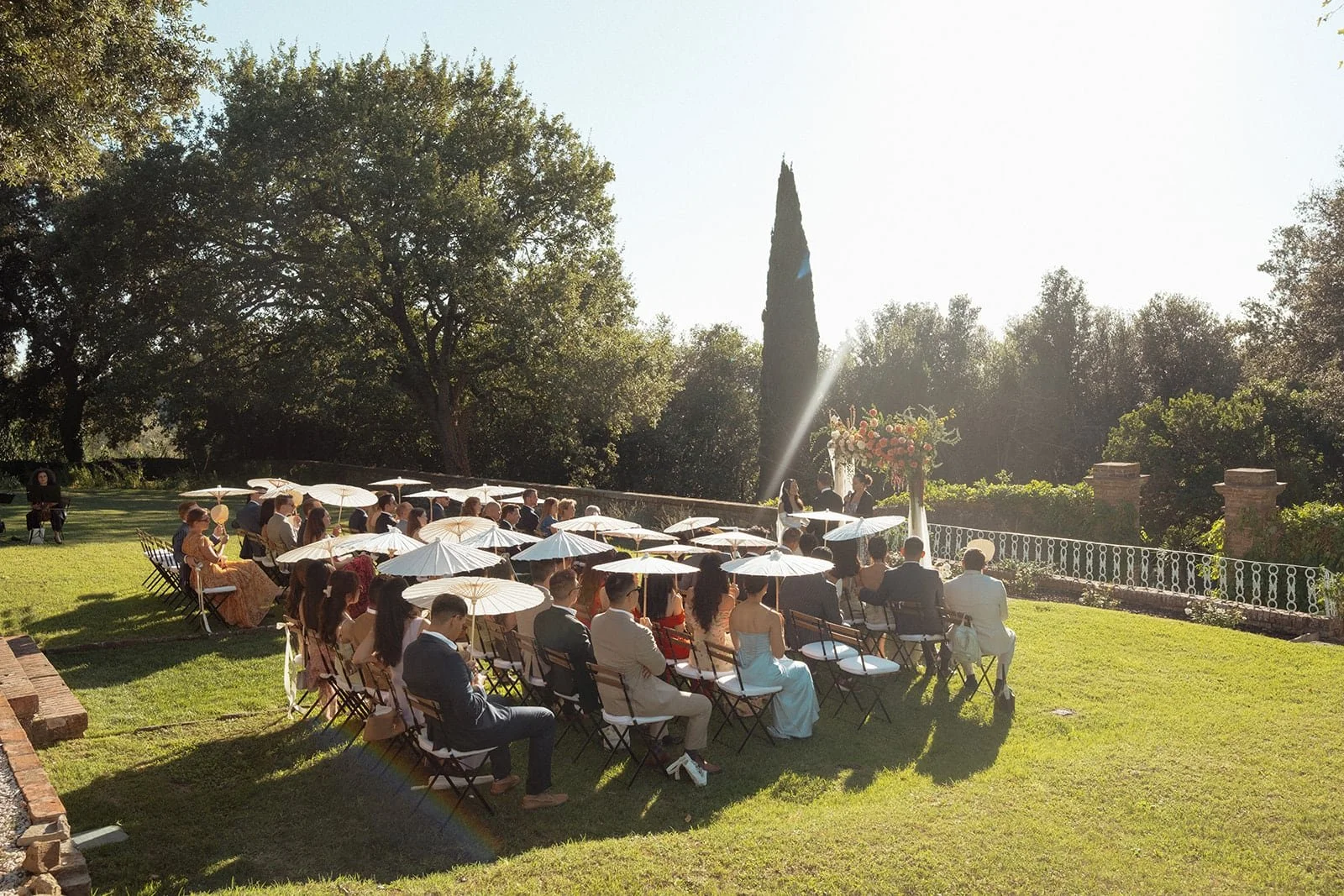 A side perspective of the ceremony at Villa Lena, captured in warm afternoon light with guests gathered beneath white parasols.