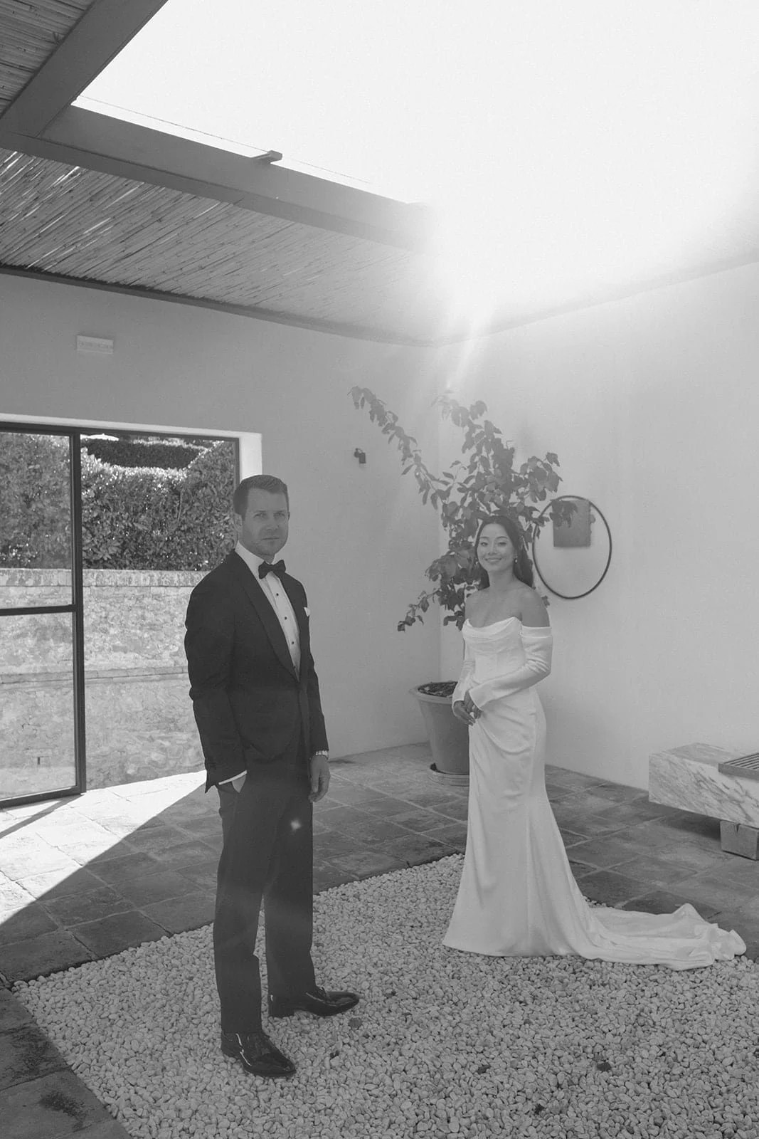 A wide black and white view of the couple standing together during their wedding ceremony, framed by natural light, shadows, and contemporary Tuscan design.