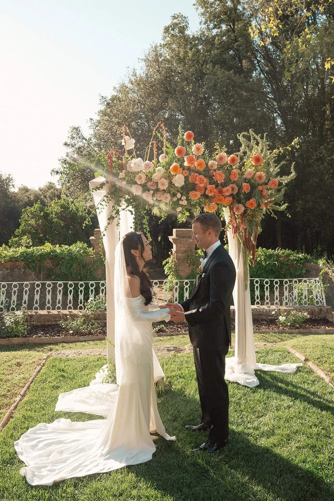 The bride and groom face each other beneath a floral arch as they exchange vows during their outdoor ceremony at Villa Lena.