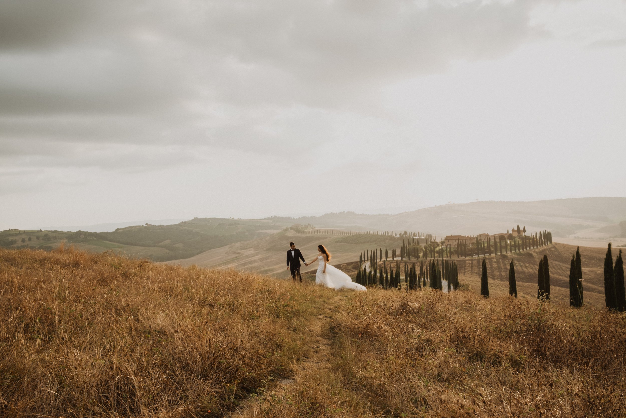 A romantic wedding moment in Val d’Orcia, Tuscany, with a couple walking through golden hills and cypress-lined landscapes, captured in soft, natural light with an editorial, timeless feel.