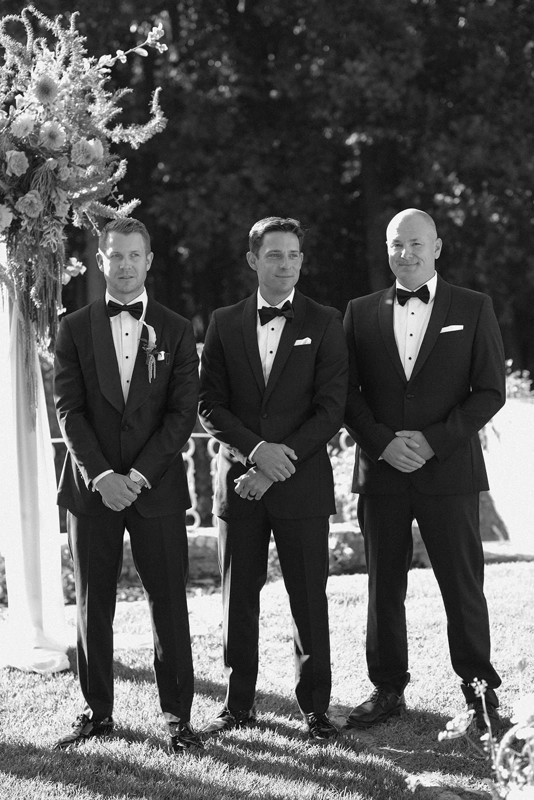 Three groomsmen stand together during the ceremony, photographed in black and white with soft light and floral details in the background.