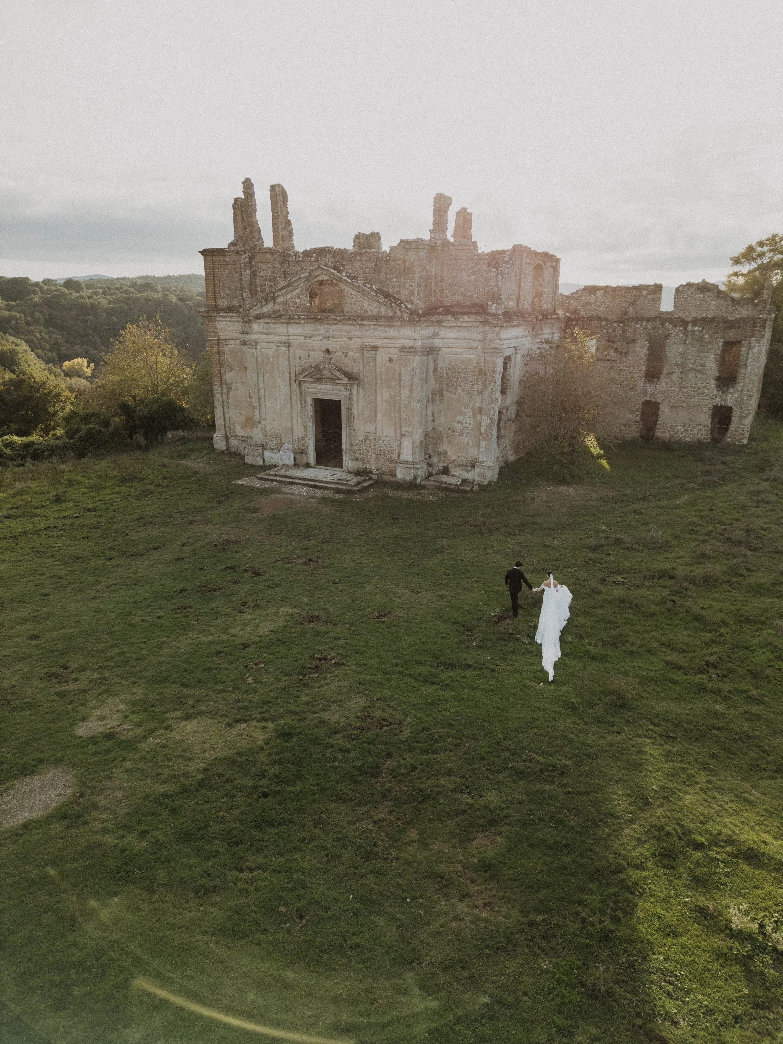 A bride and groom walk hand in hand toward a historic Italian ruin, captured in soft natural light. A poetic, documentary wedding photograph that reflects intimacy, landscape, and a refined sense of place in Italy.