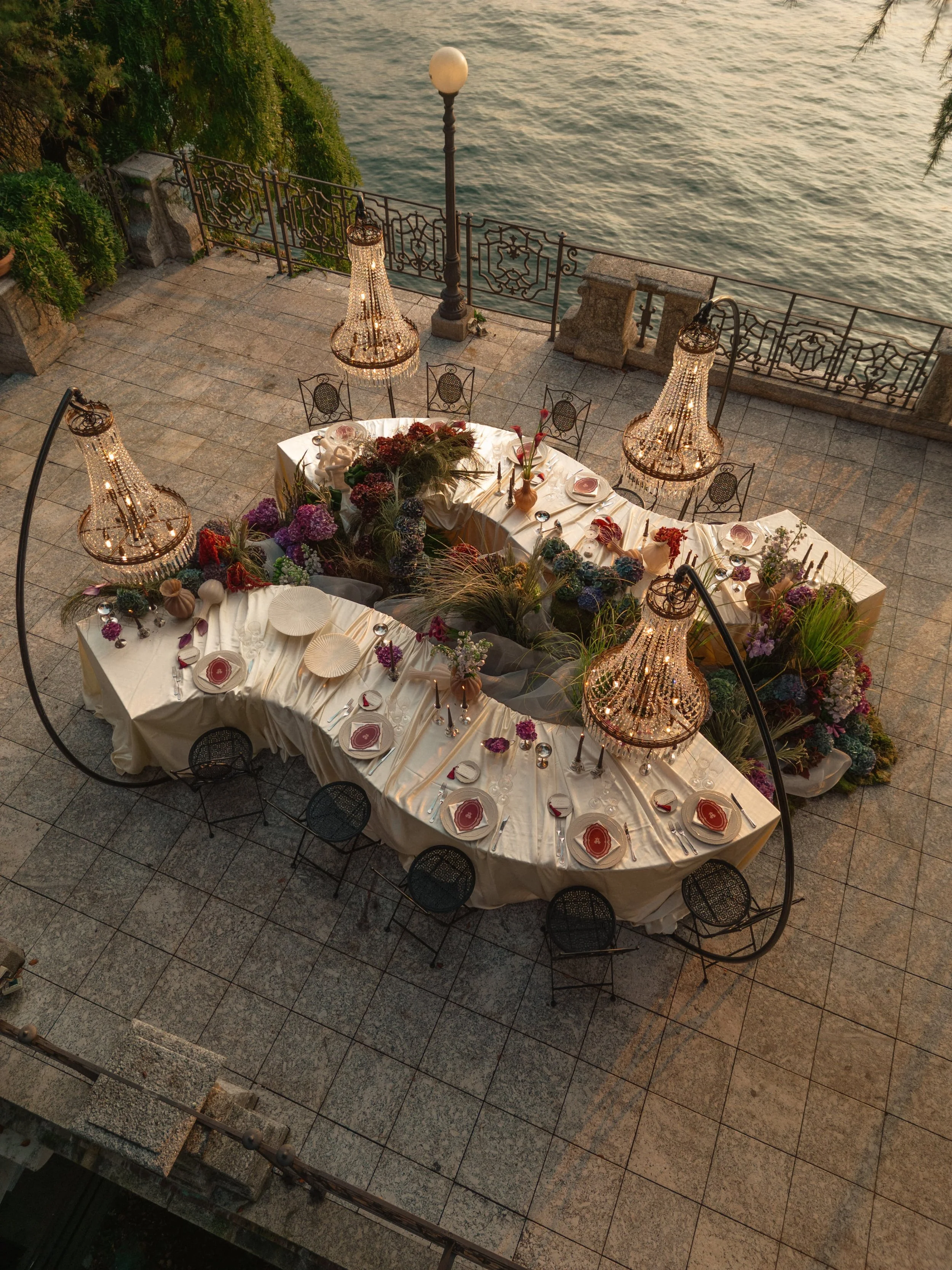 An intimate luxury wedding dinner set on the shores of Lake Como, featuring sculptural floral design, candlelight chandeliers, and an elegant tablescape overlooking the water at sunset.