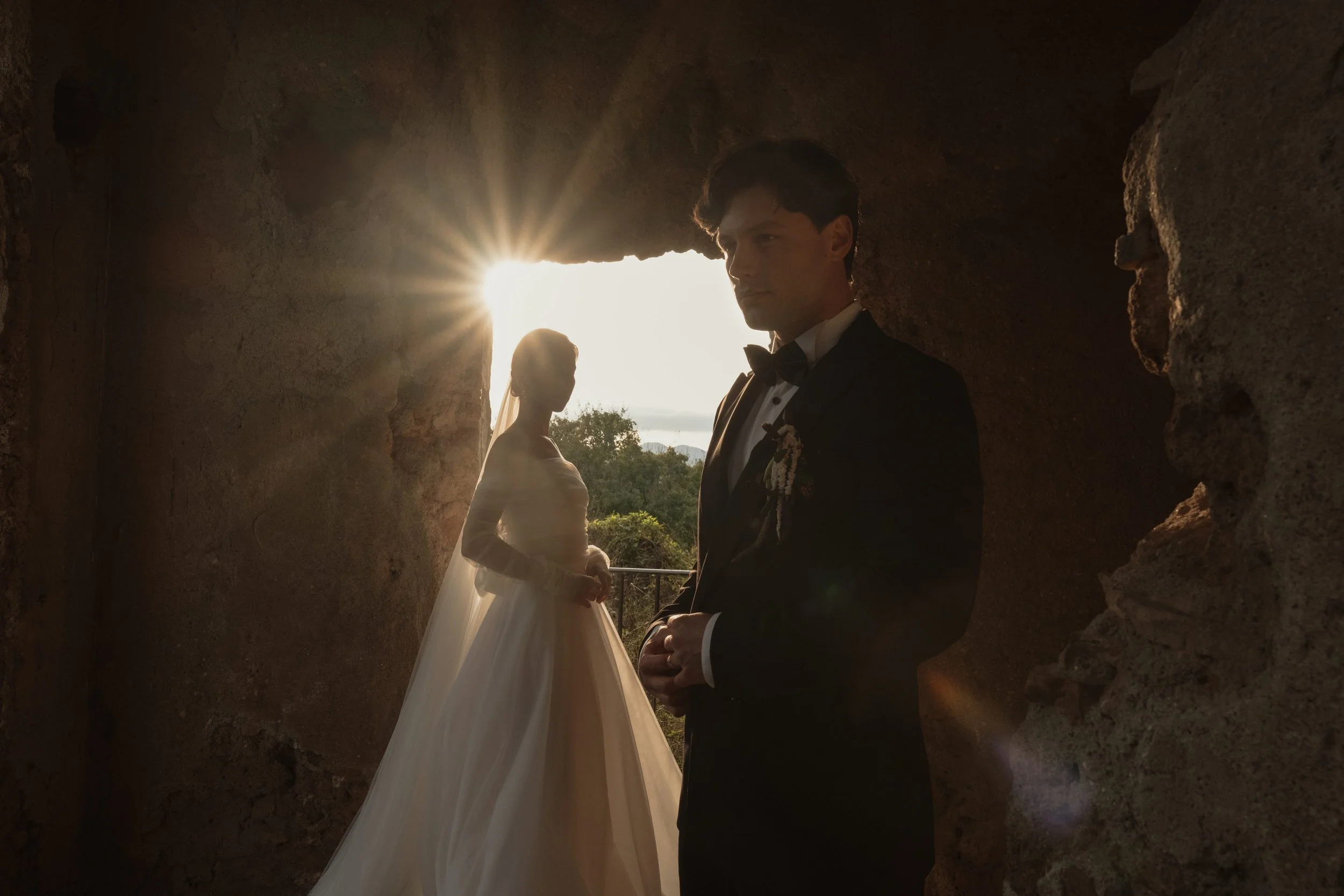 A bride and groom framed by ancient stone walls in Italy as golden light filters through the ruins. A cinematic, documentary wedding portrait in Italy, capturing intimacy, atmosphere, and timeless elegance through natural light and shadow.